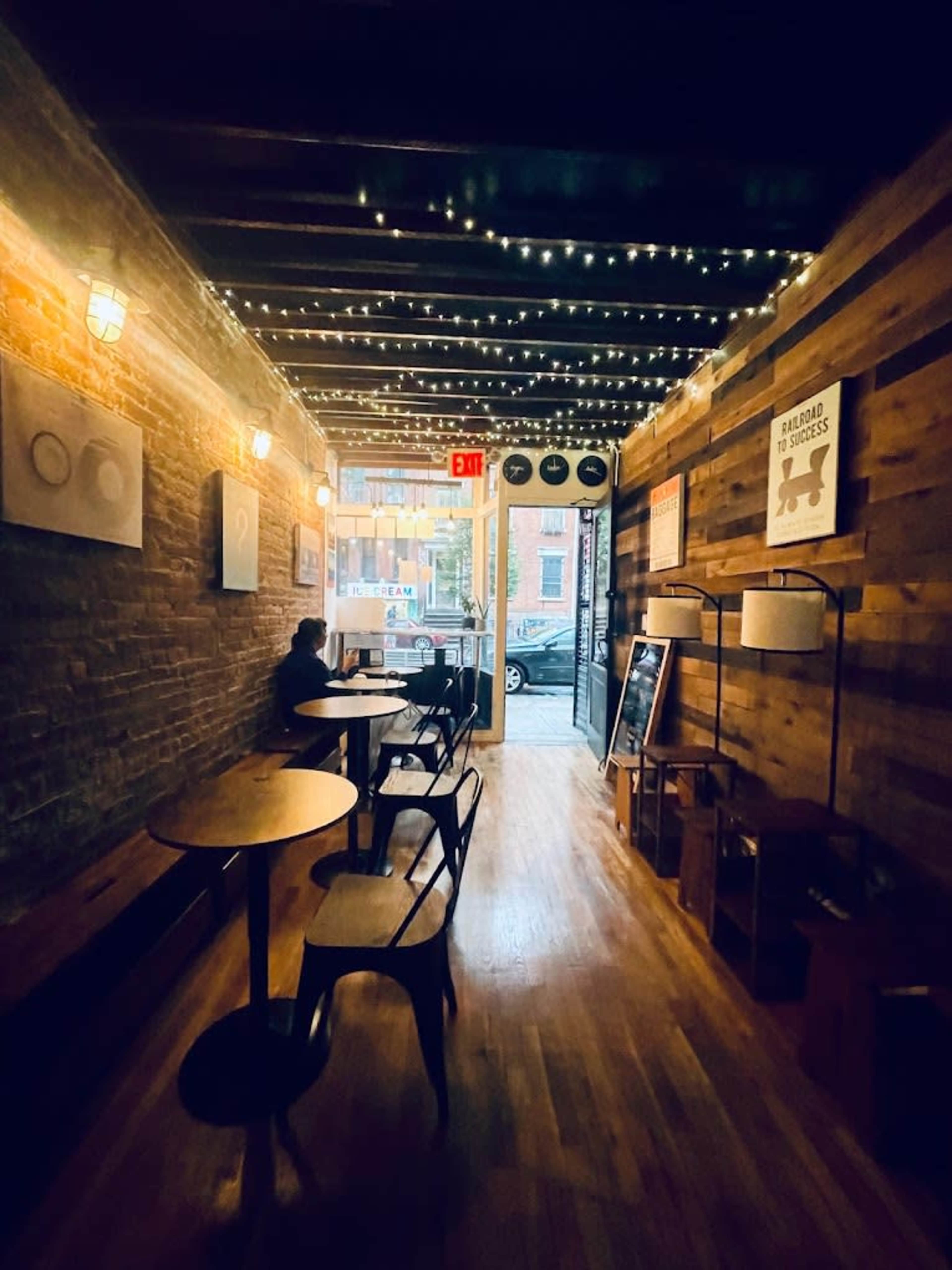 A dimly lit hallway of a café with wooden walls, small tables, and string lights overhead, leading to an exit that opens to the street.