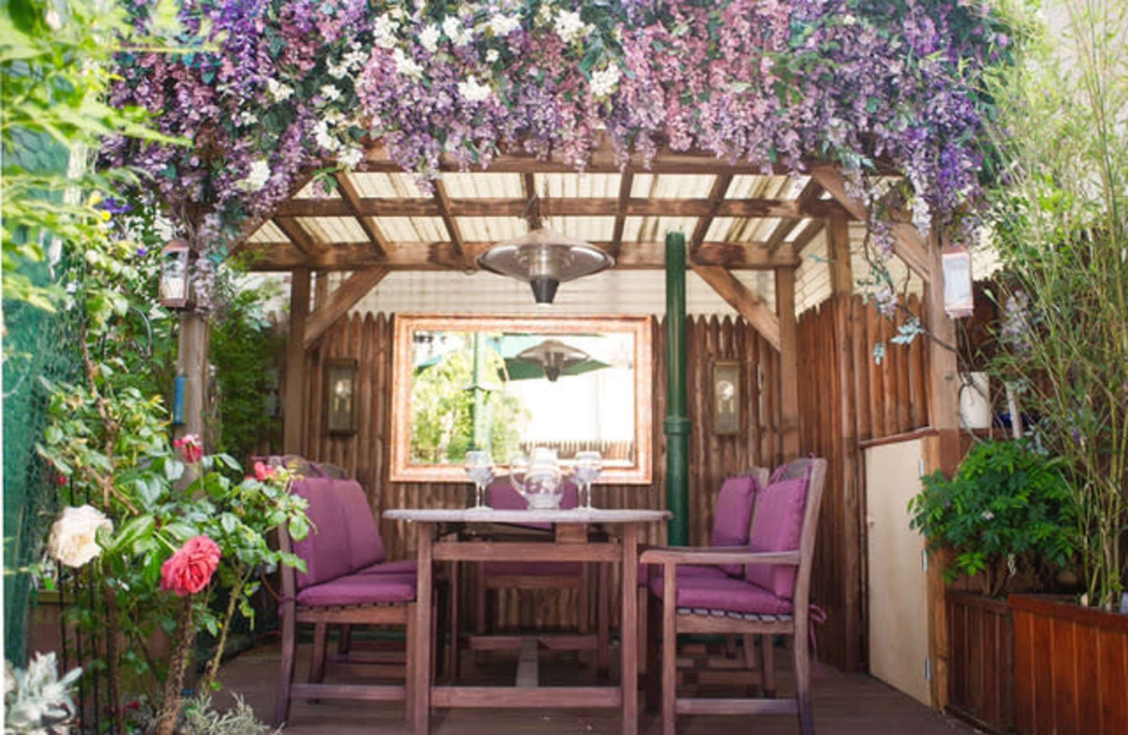 A cozy outdoor seating area adorned with purple flowers, featuring a wooden table and chairs under a decorative canopy.