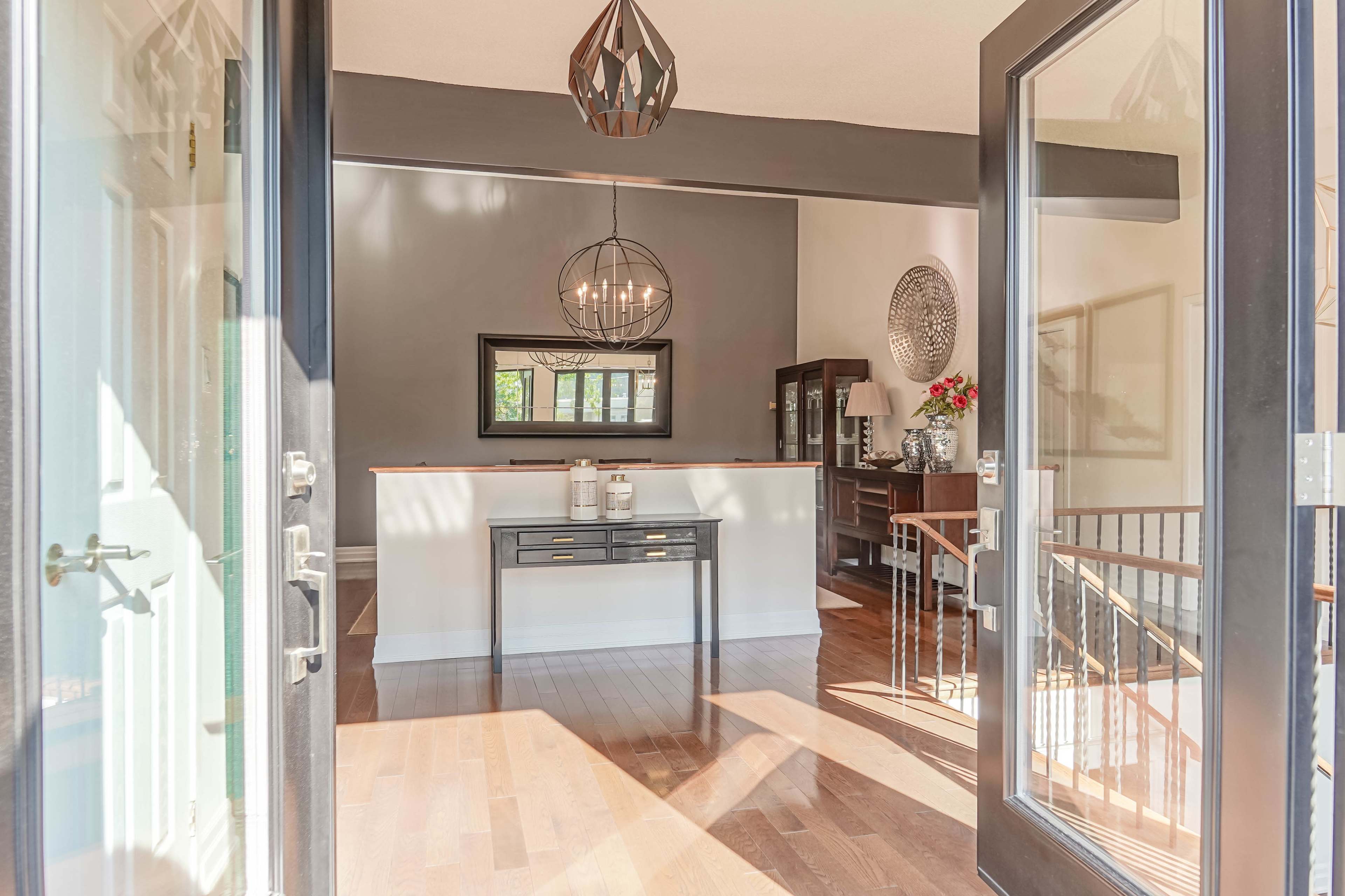The image shows a welcoming entryway with a table, a mirror, and a dining area illuminated by natural light streaming through large doors.
