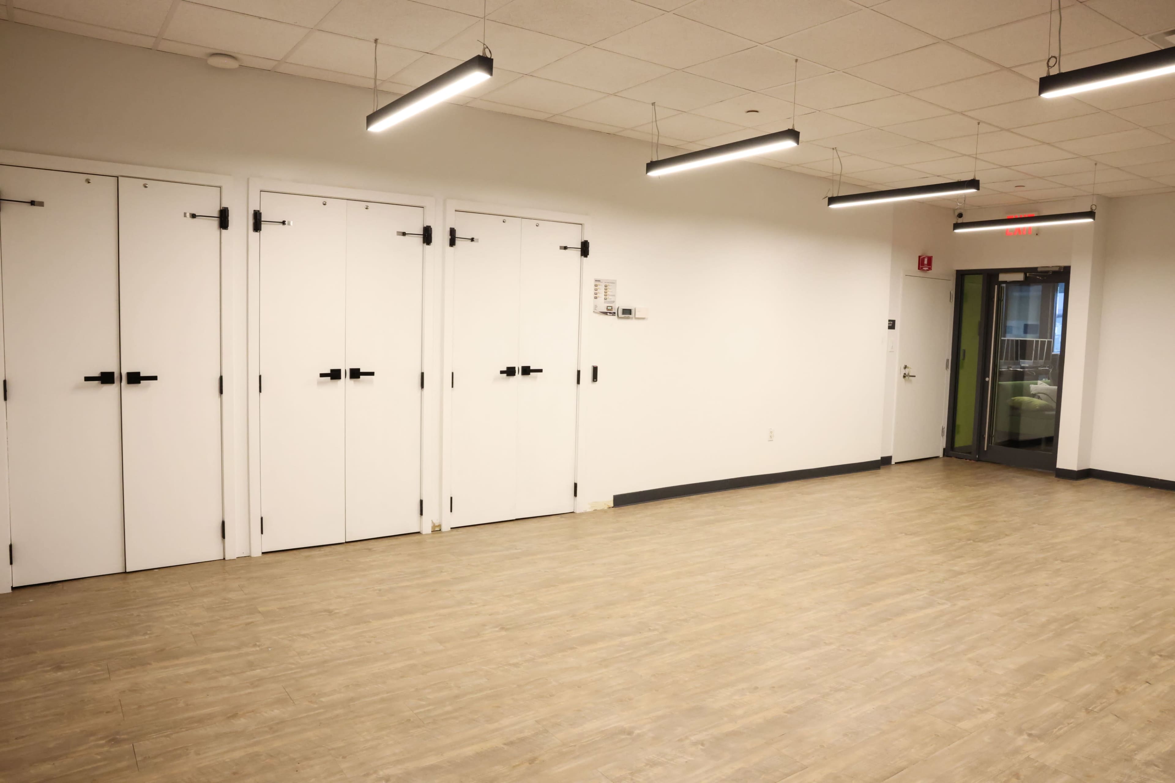 The image shows an empty room with pale wooden flooring, white walls, and three closed doors on one side.