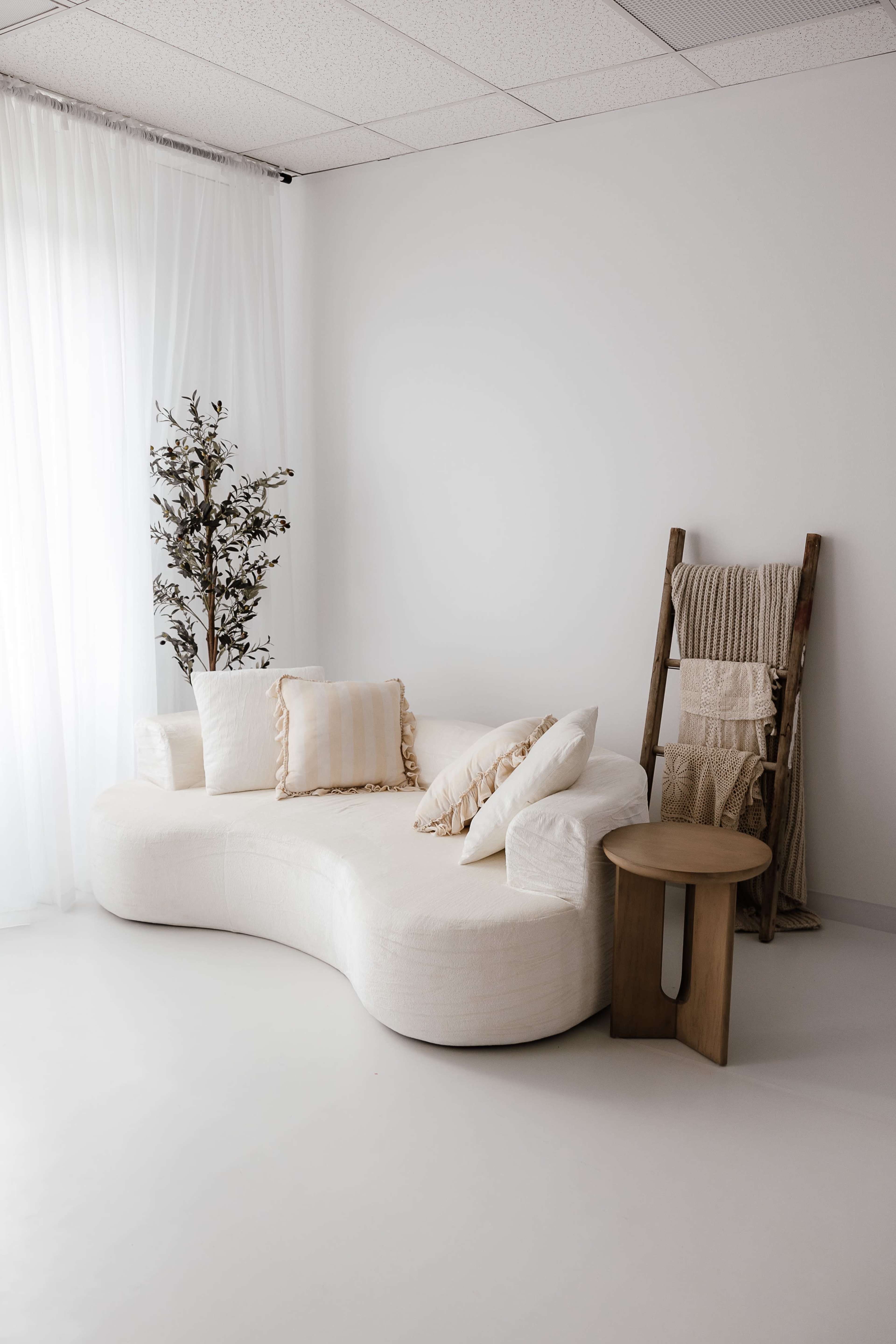 A curved white sofa with several decorative pillows is positioned next to a small wooden side table and a plant, against a minimalist backdrop of white walls and sheer curtains.