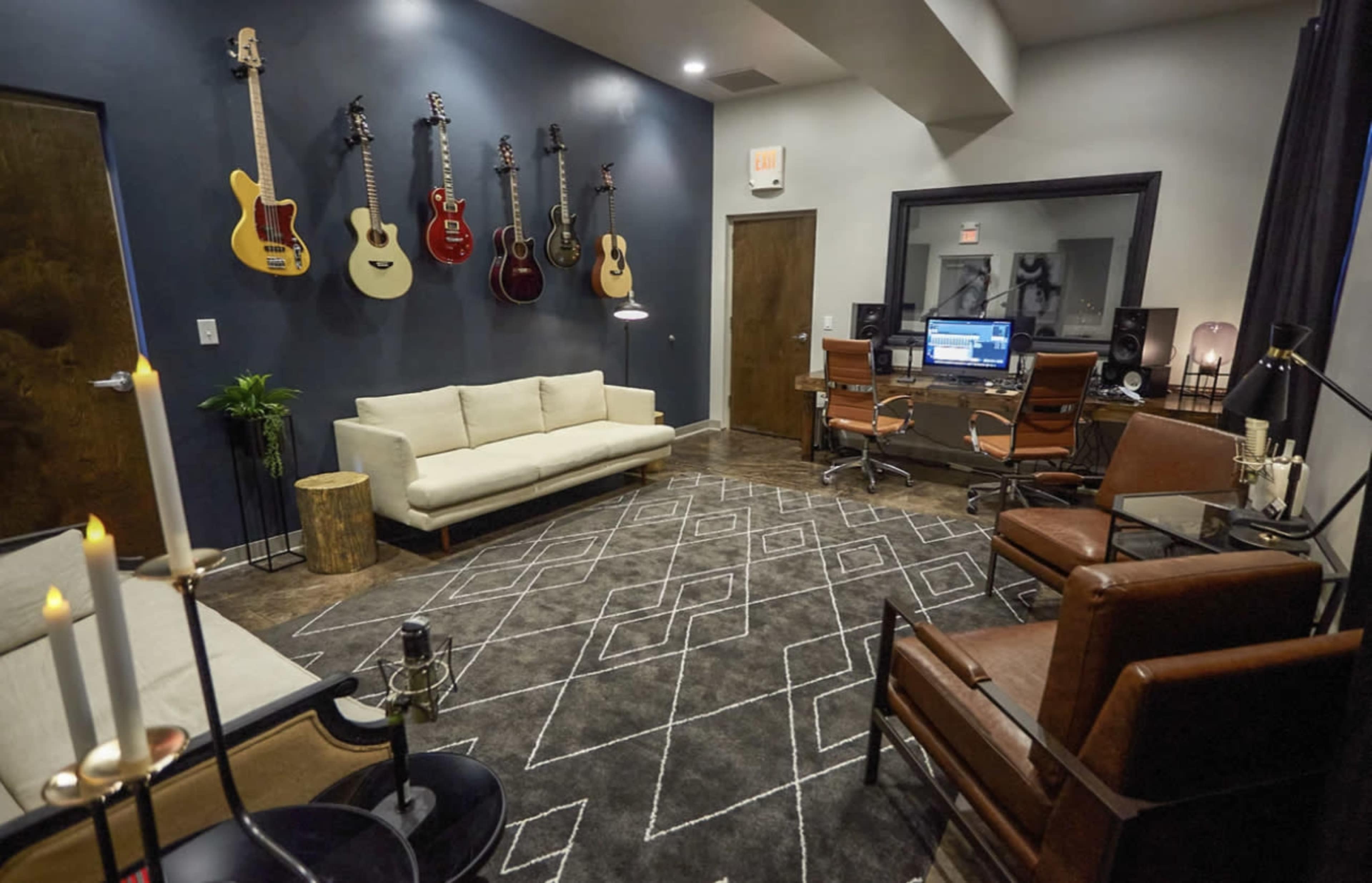 A modern music studio features a couch, two armchairs, and several guitars hanging on the wall, with a workspace containing audio equipment visible in the background.