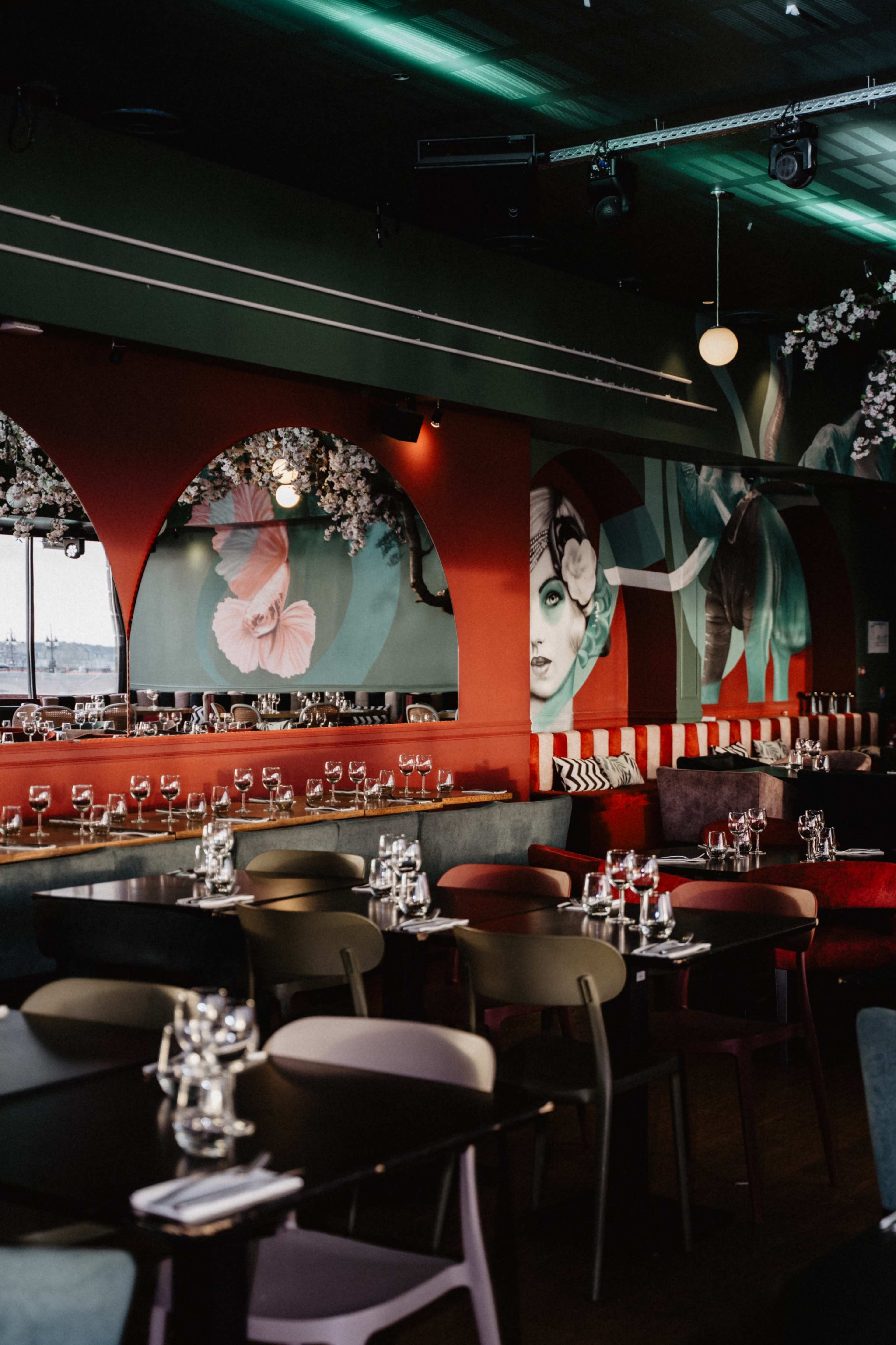 The image shows a colorful restaurant interior featuring arched windows, artistic wall murals, and tables set with glassware and chairs of various colors.