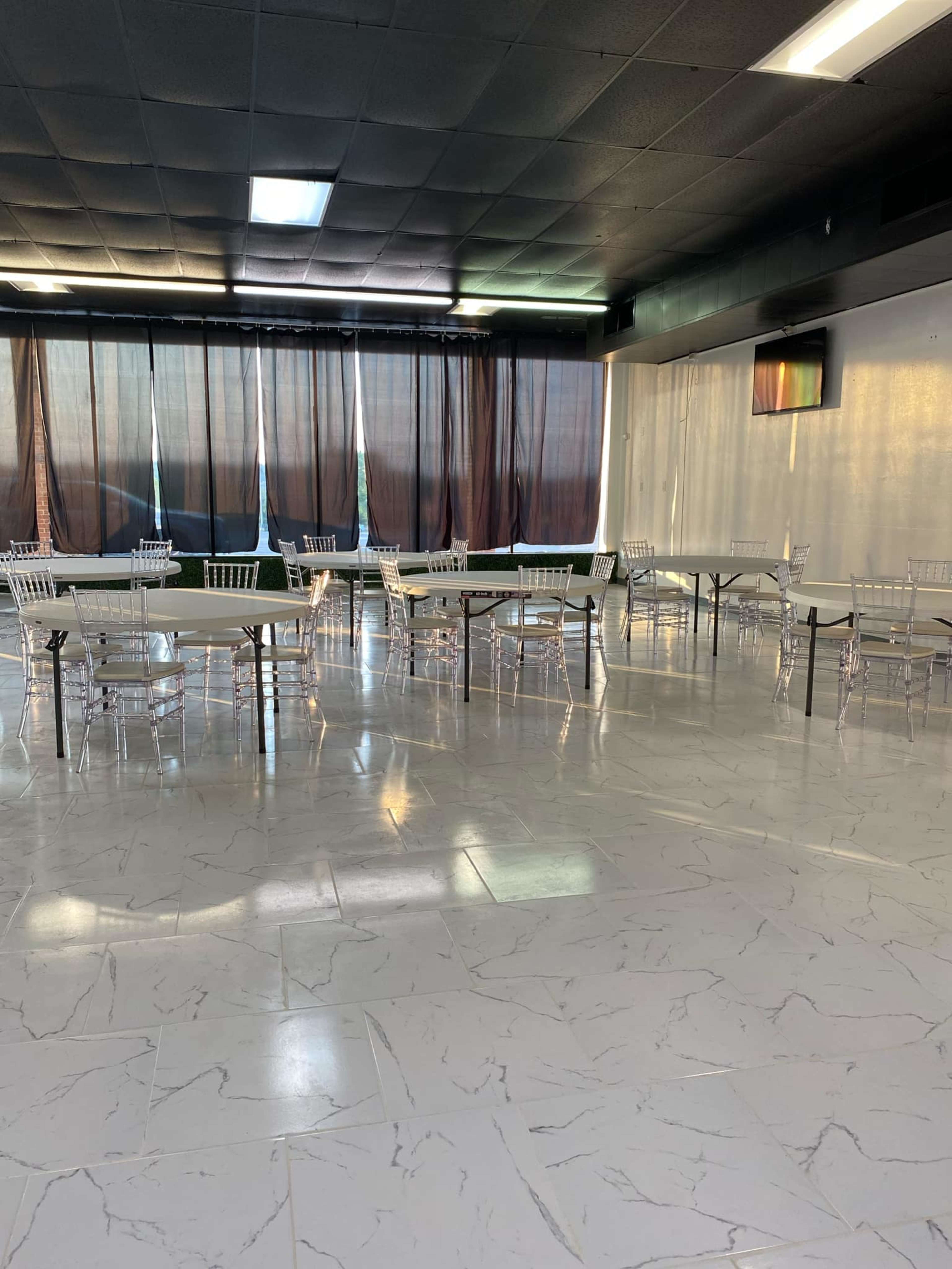 A spacious, empty event room with several tables and chairs set up, illuminated by natural light coming through large black curtains.