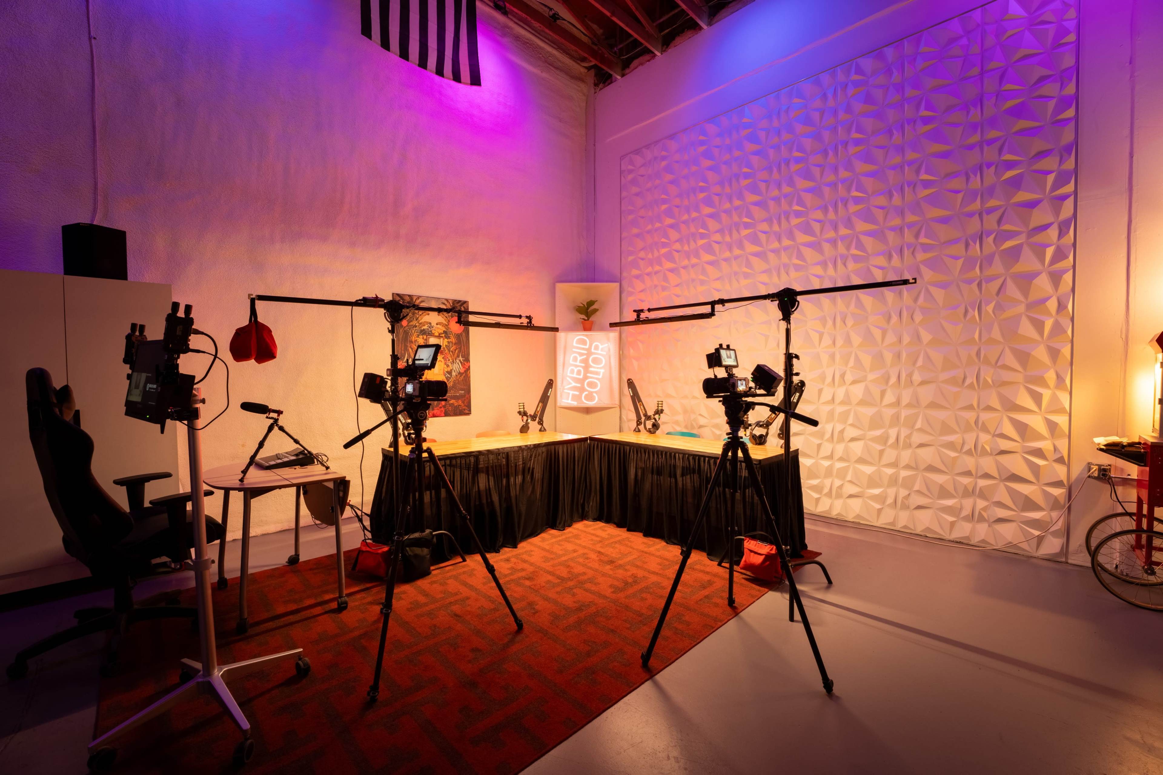 A video production studio equipped with two cameras on tripods, a desk with lighting equipment, and a textured backdrop illuminated in purple hues.