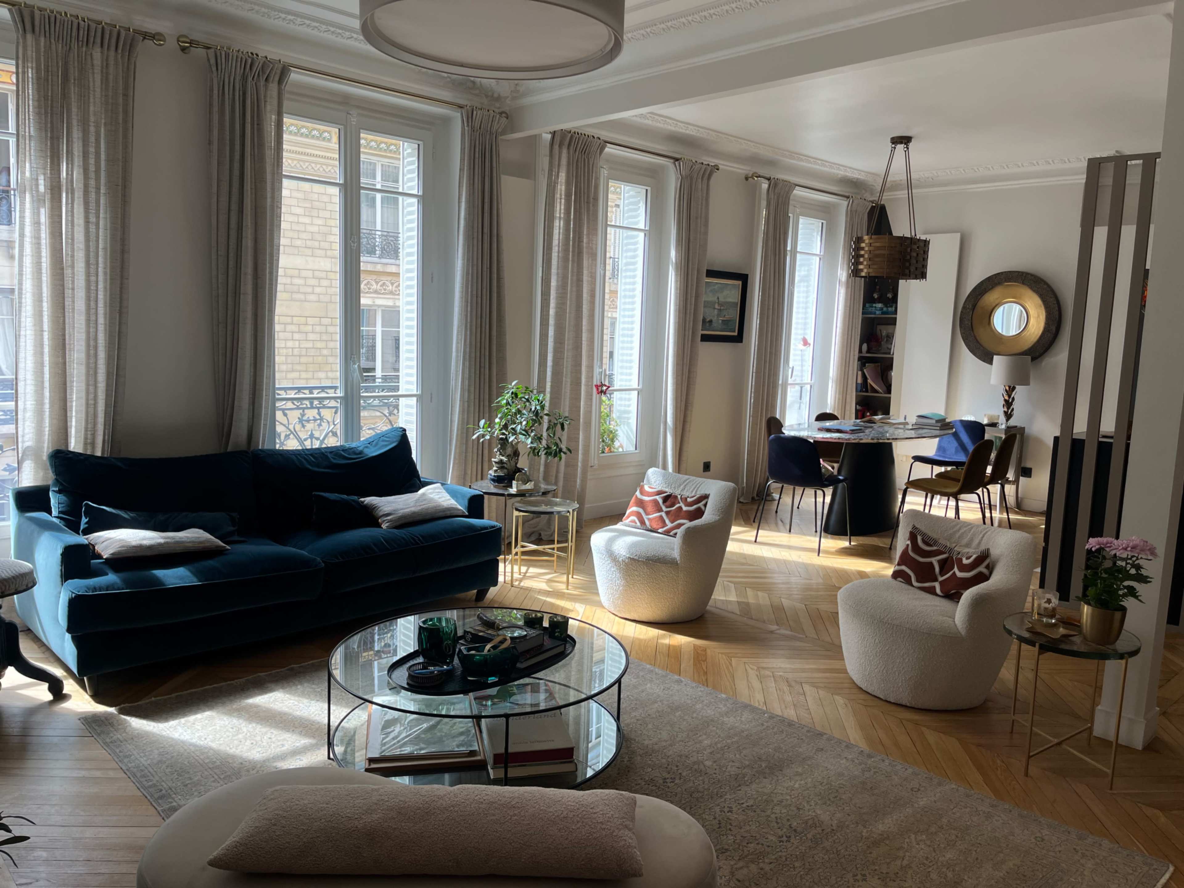 The image shows a sunlit living room featuring a blue sofa, two white armchairs, and a glass coffee table, with a dining area visible in the background.