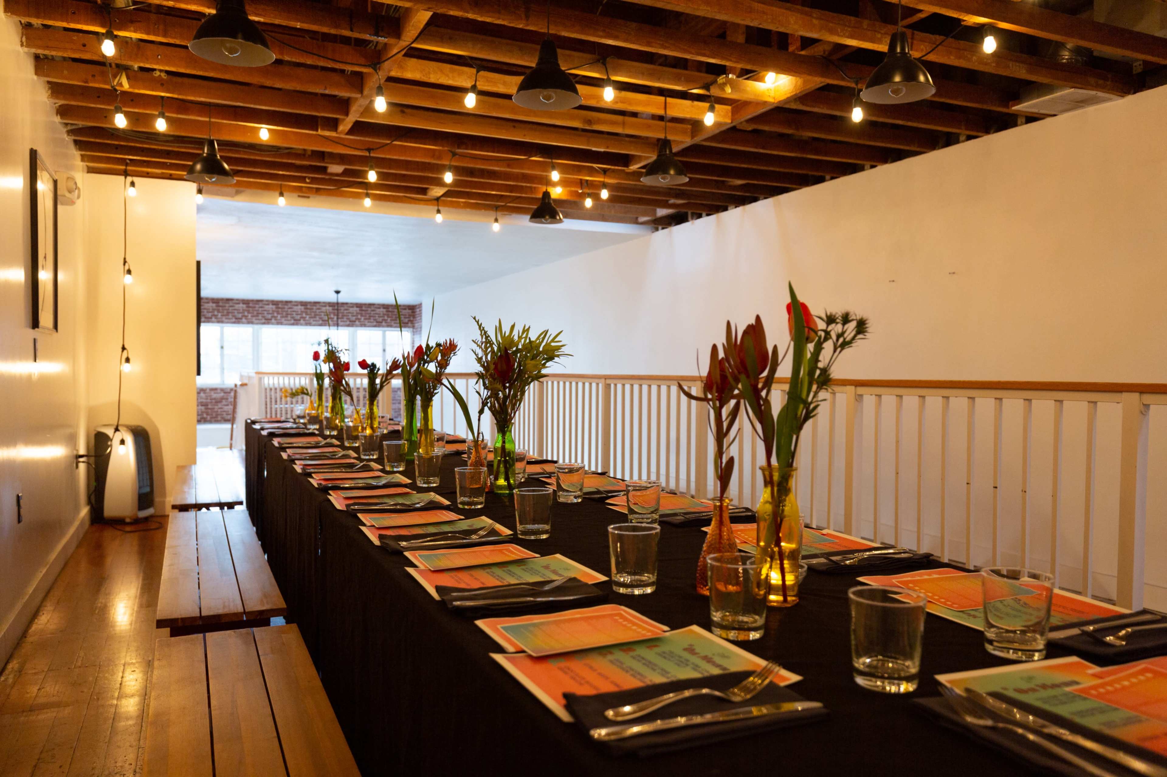 A long dining table is set with colorful menus and vases of flowers in a well-lit, industrial-style space.