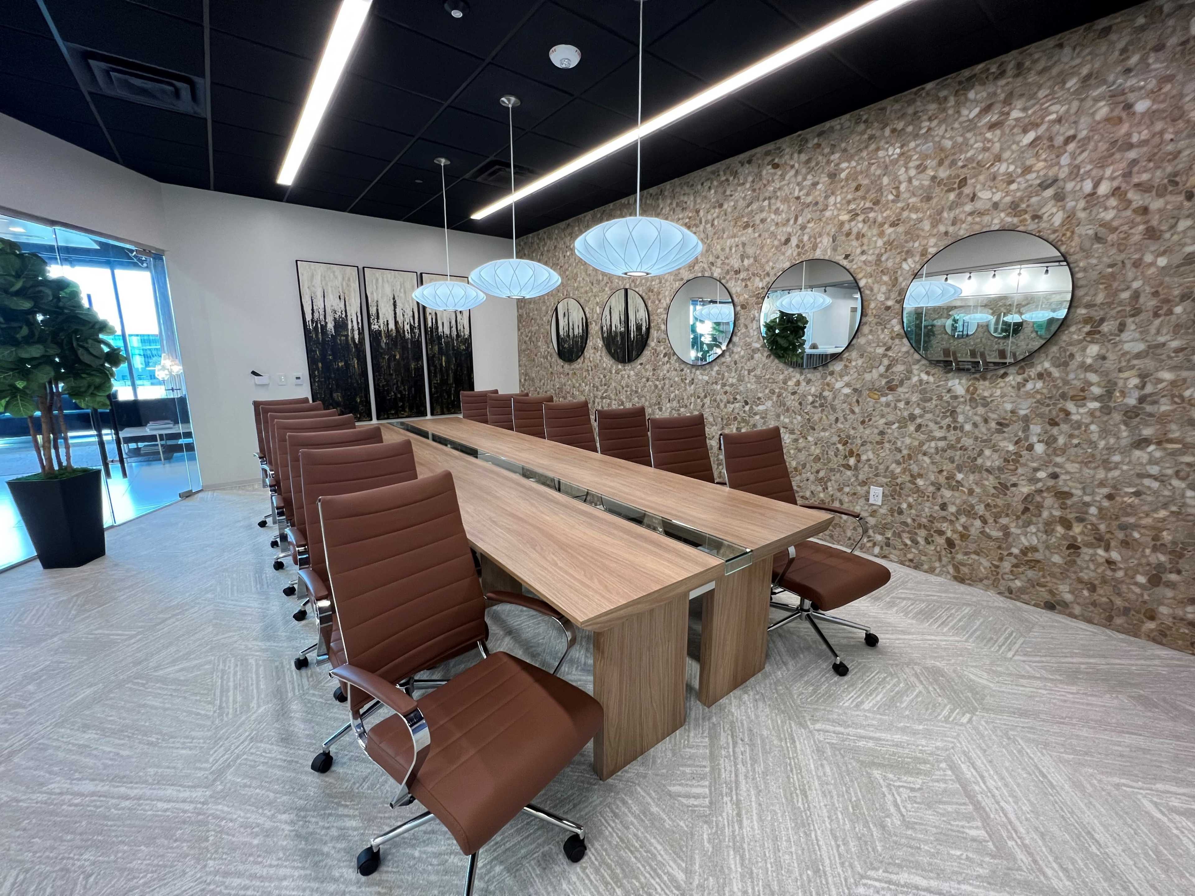 A modern conference room with a long wooden table surrounded by brown office chairs, featuring a textured stone wall and pendant lights above.