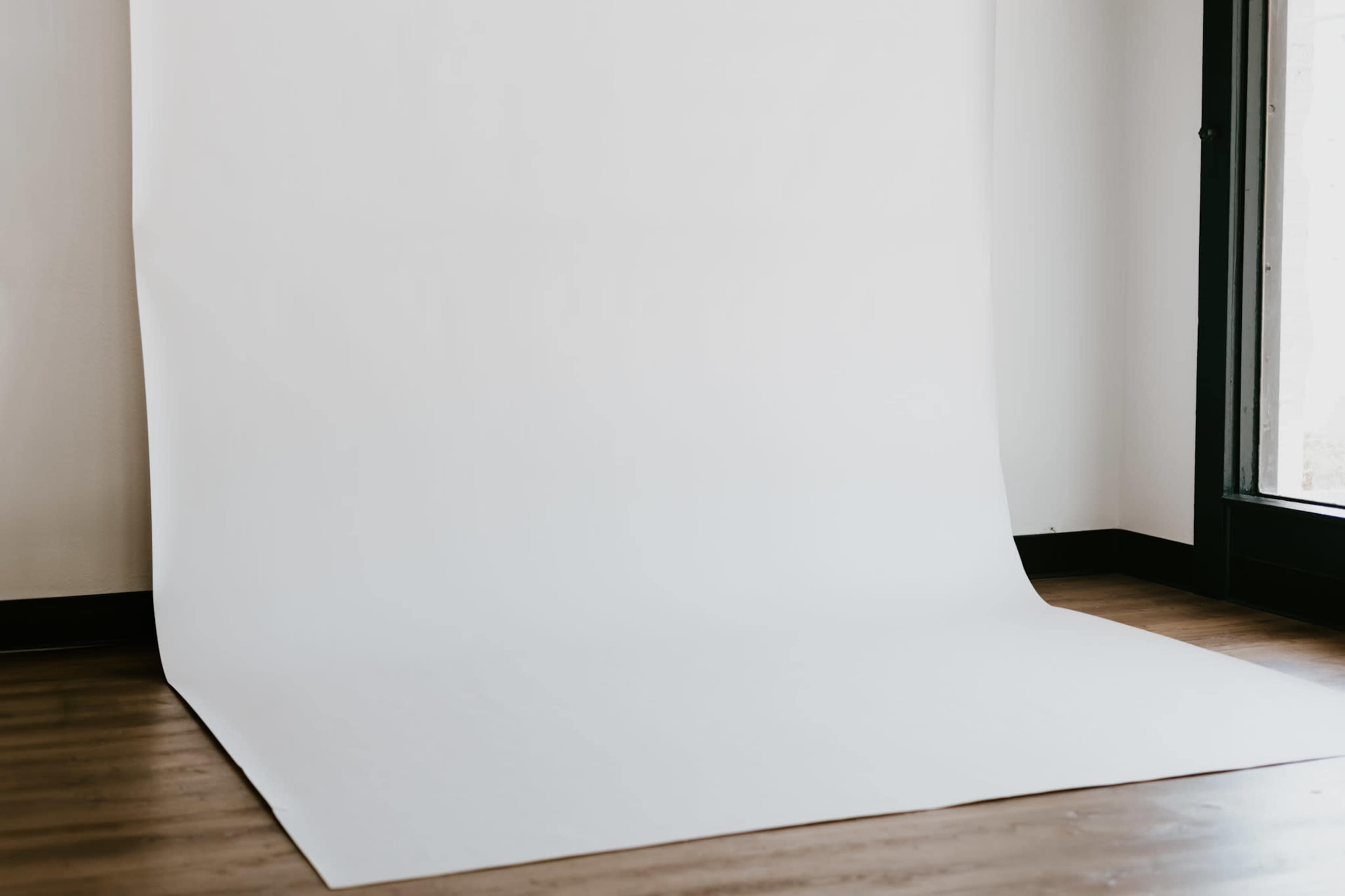 The image shows a blank white backdrop set up against a wall in a room with wooden flooring and a window.