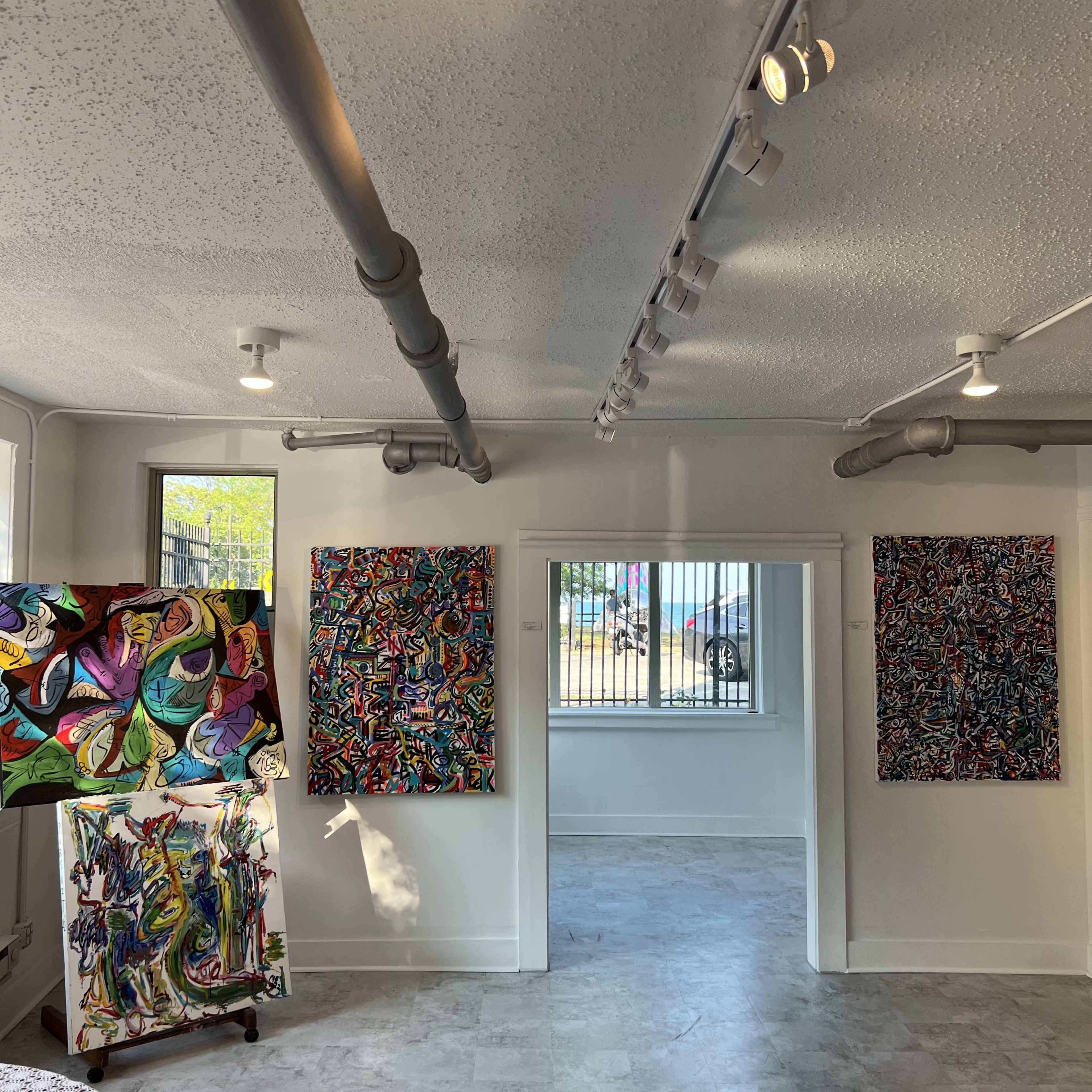 The image shows an art gallery featuring vibrant abstract paintings on white walls, with a view of an outdoor area through a window.