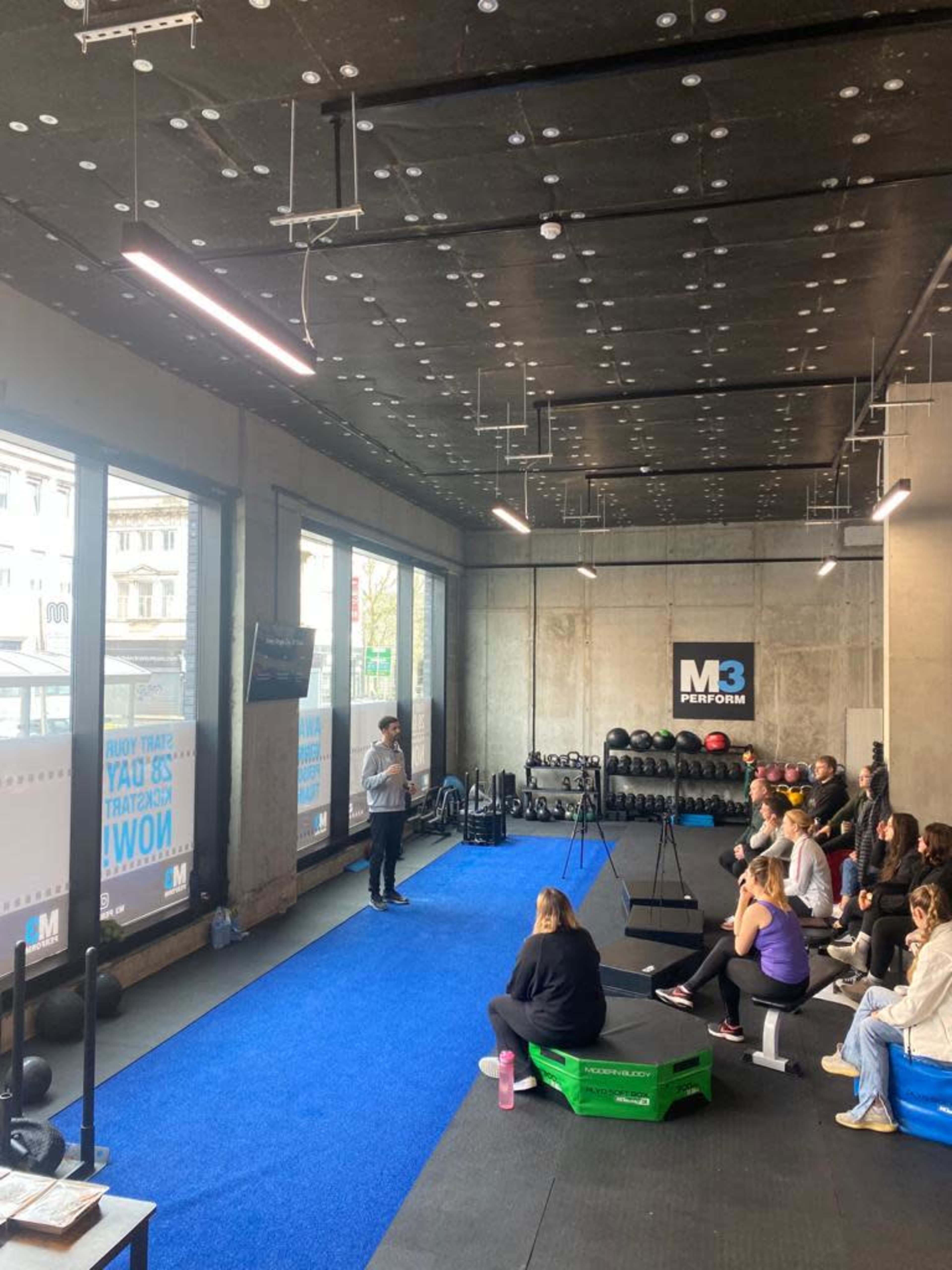 A fitness instructor is speaking to a group of people seated on a blue mat in a modern gym with large windows.