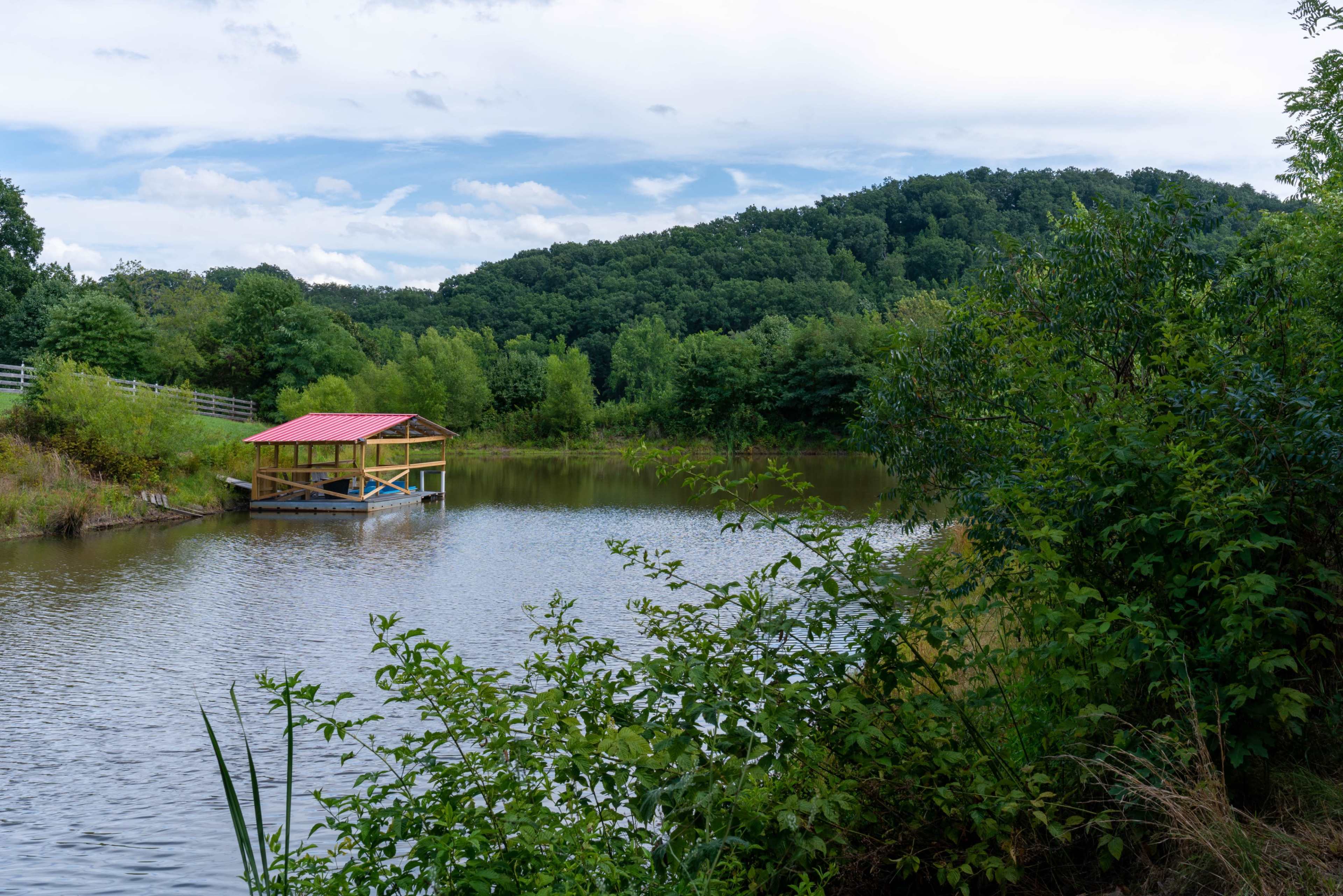 A wooden dock with a red roof is situated on a calm pond surrounded by lush greenery and rolling hills.