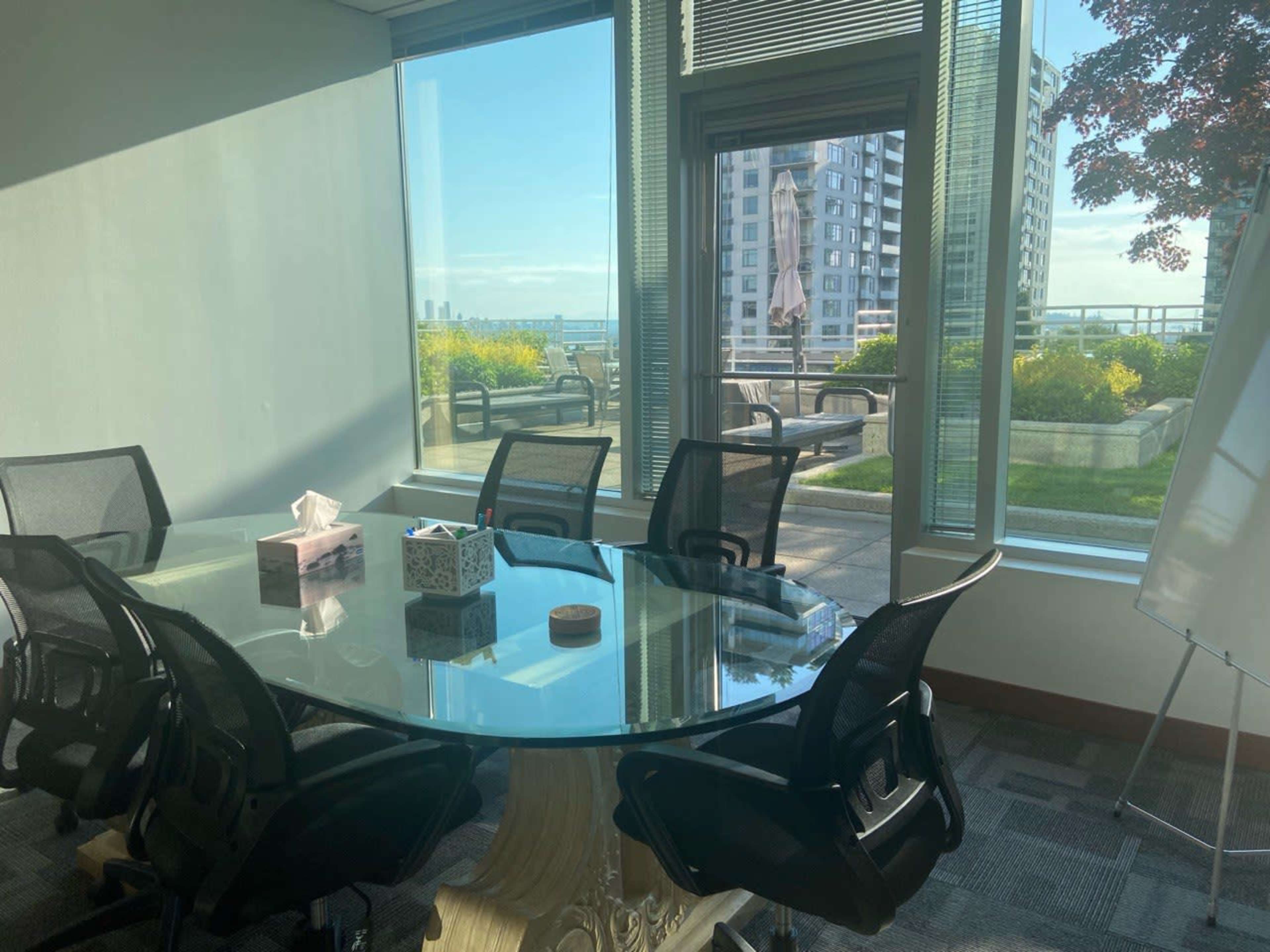 A modern conference room features a glass table surrounded by black chairs and large windows overlooking a green outdoor terrace and city skyline.