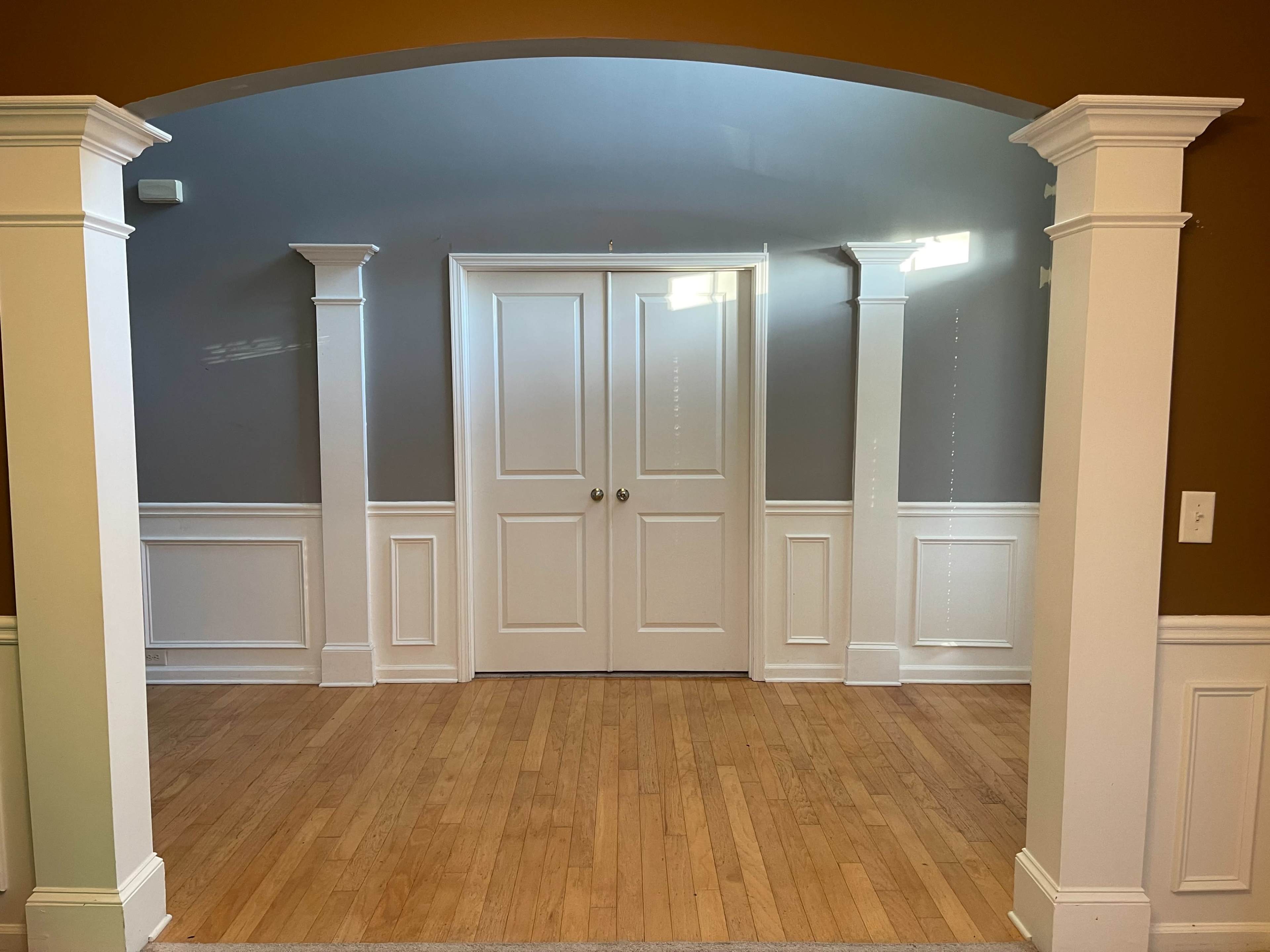 An empty room with hardwood flooring, white wainscoting, and two columns framing an entrance with double doors.