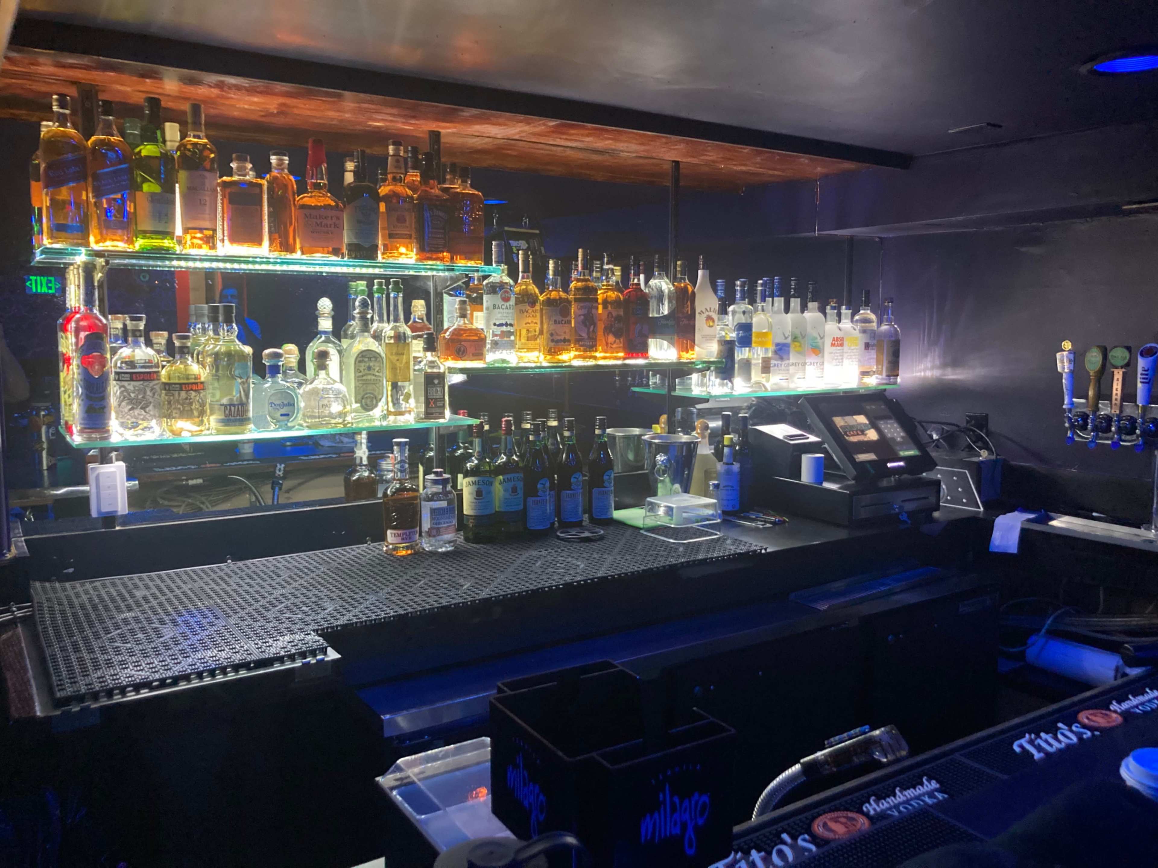 A dimly lit bar showcases shelves lined with various bottles of liquor and a well-organized countertop with glassware and taps.