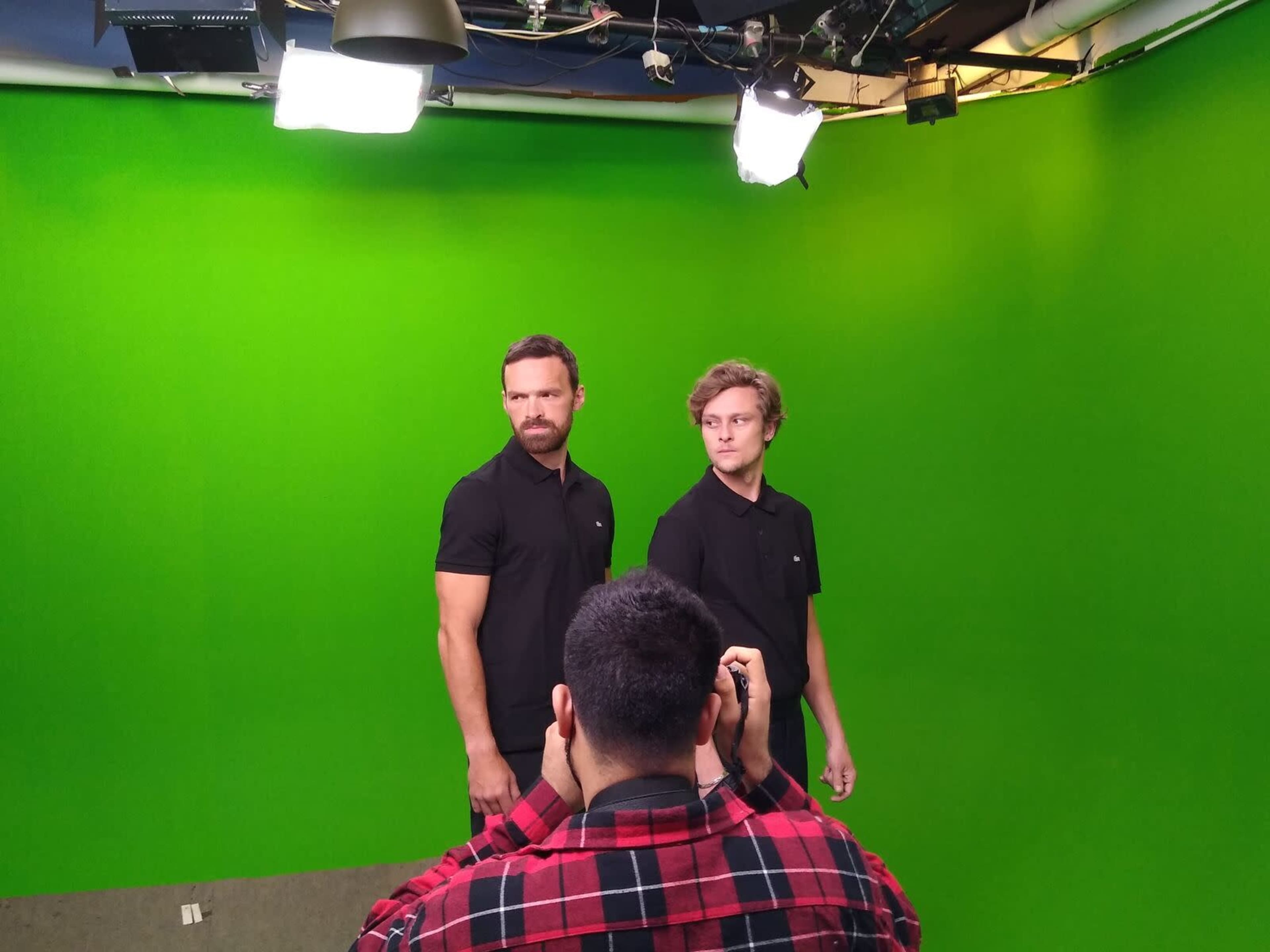 Two men in black polo shirts stand in front of a green screen while a third person takes their photo.