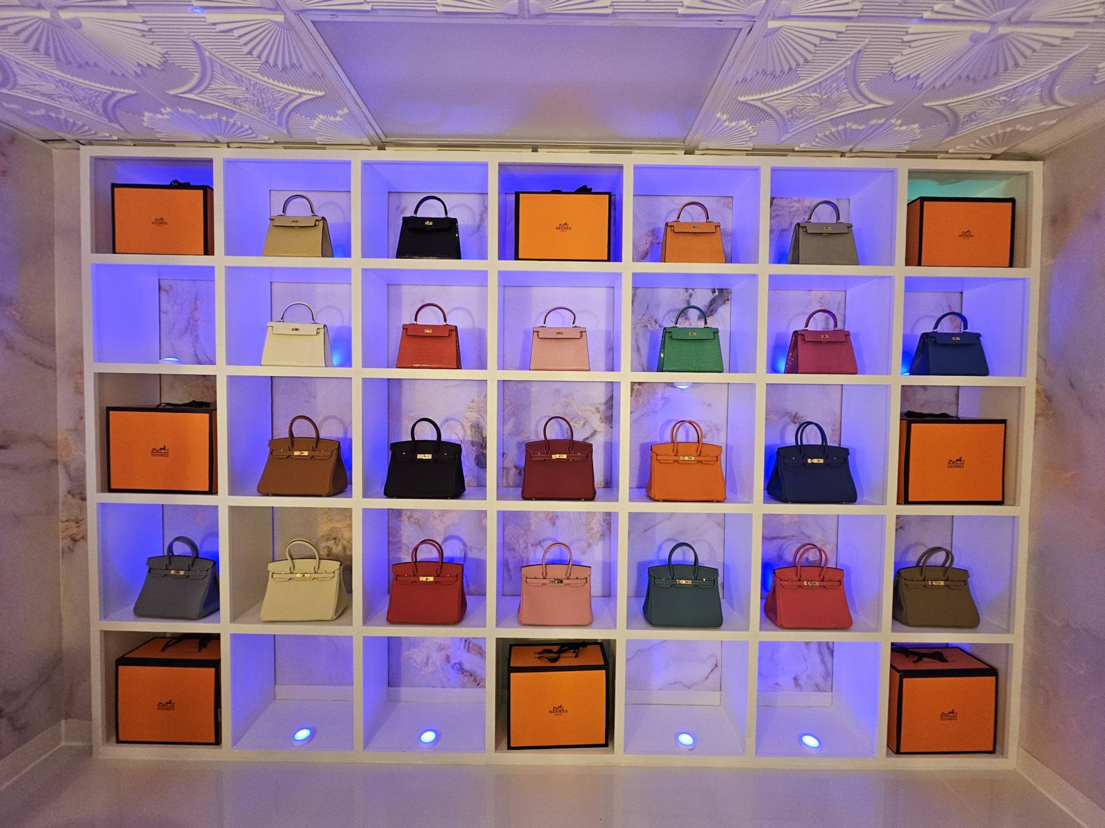 A display wall with organized shelves showcasing various handbags against a marble background, illuminated by blue lighting.