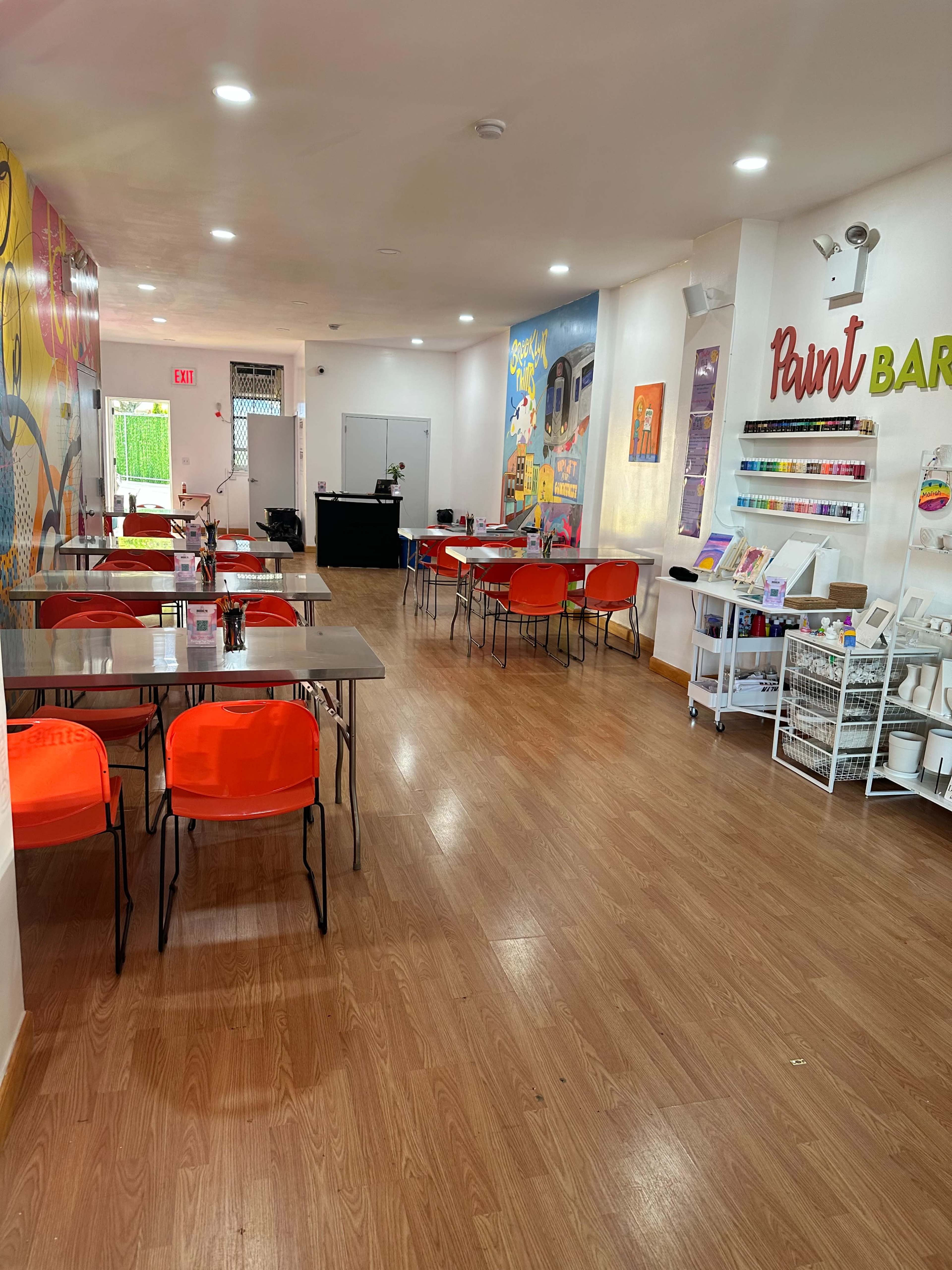A brightly lit art studio featuring several tables with orange chairs, a wall mural, and shelves displaying art supplies.