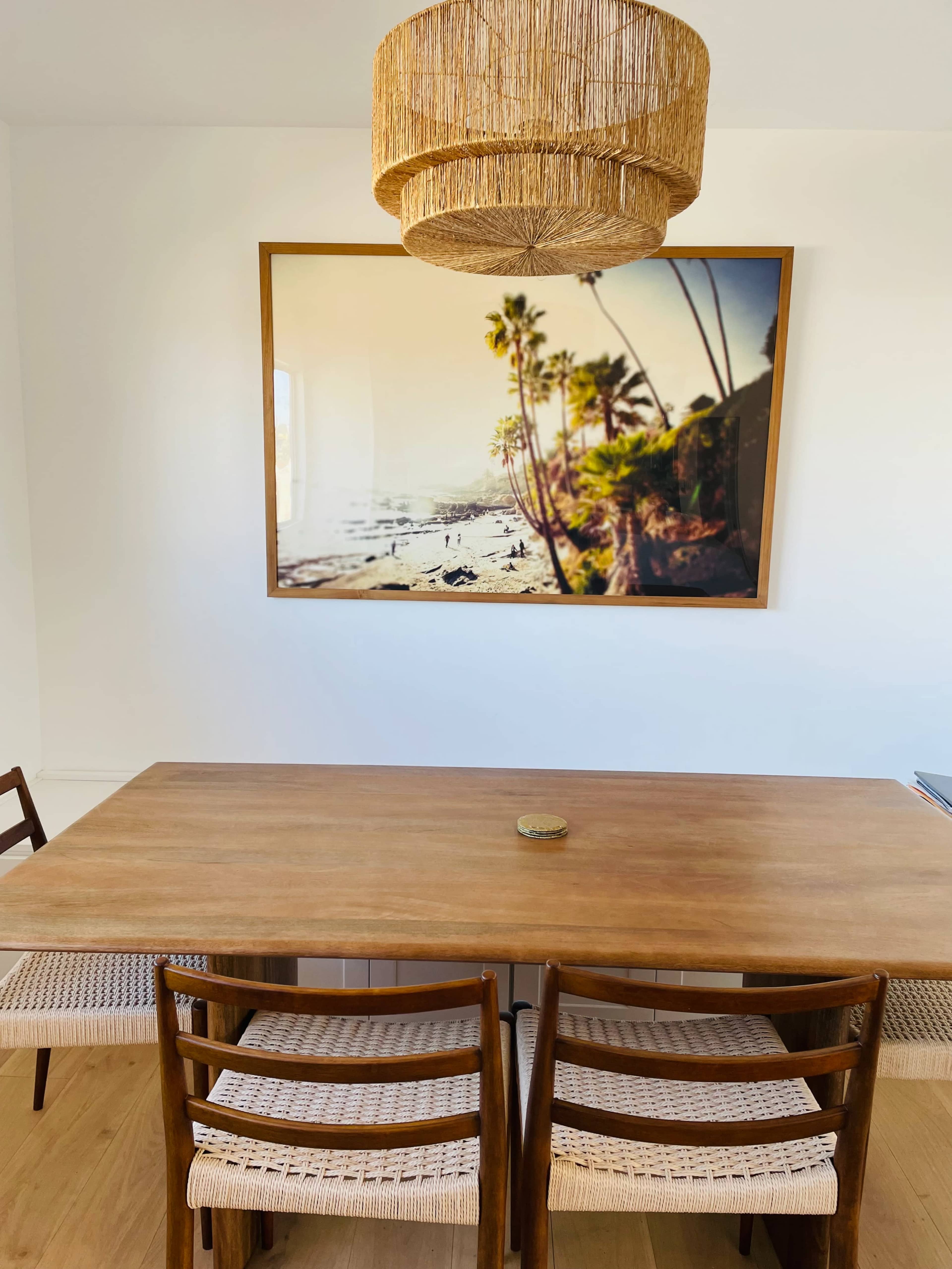 A wooden dining table with woven chairs is set beneath a straw pendant light and a large framed photograph of a coastal scene featuring palm trees.