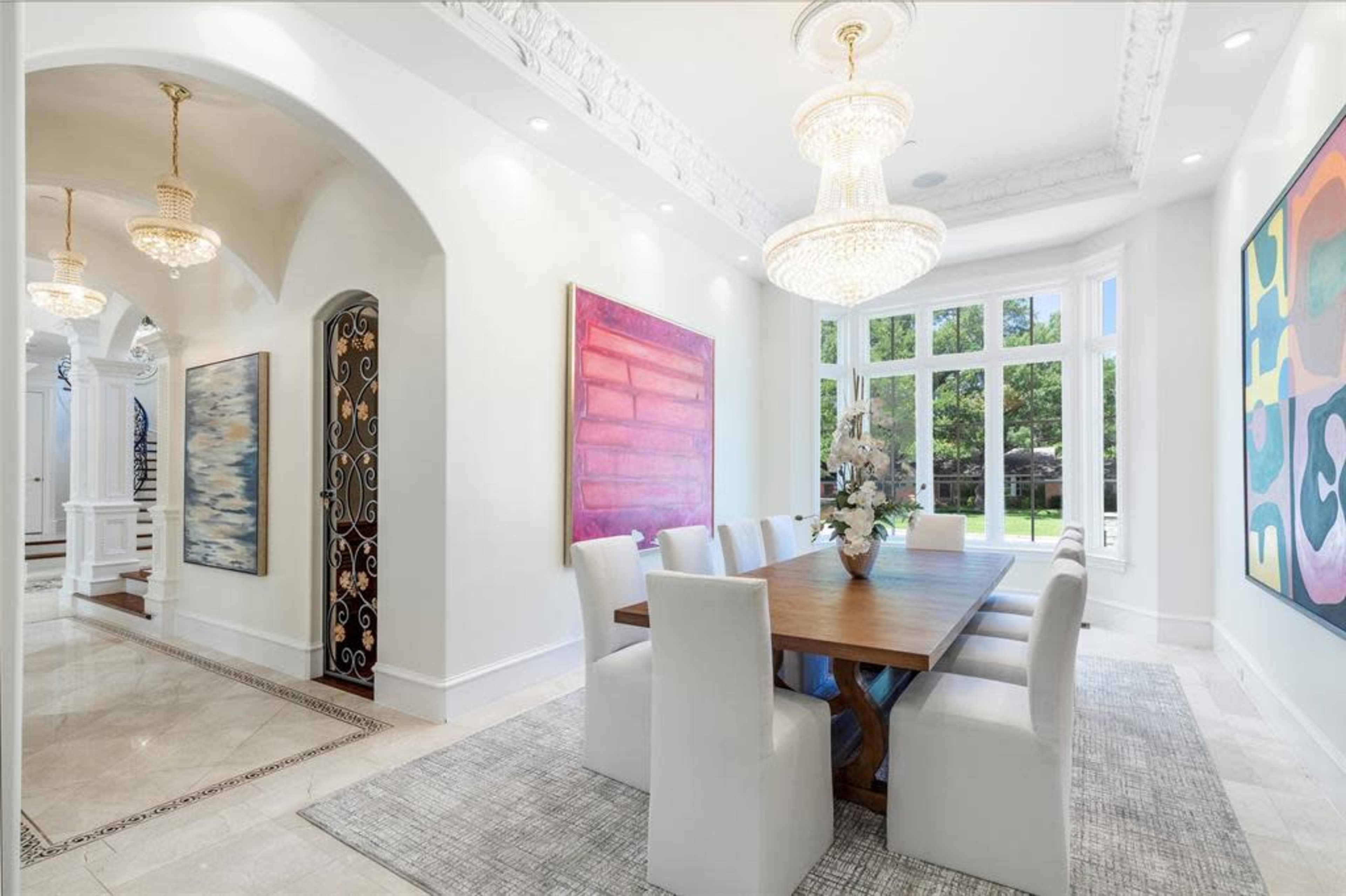 The image shows a modern dining room with a long wooden table surrounded by white chairs, featuring large windows and colorful artwork on the walls.