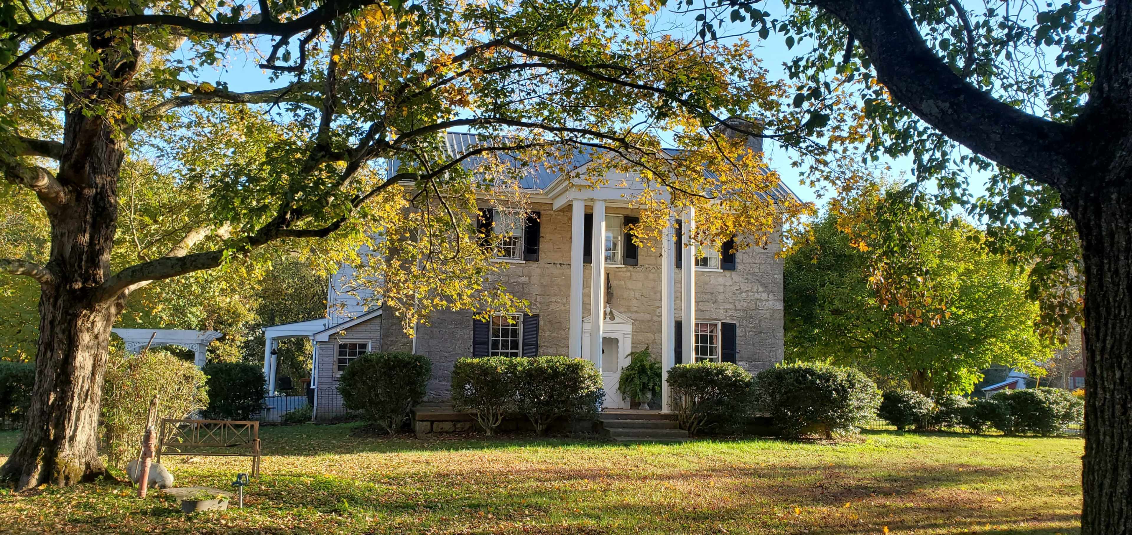 A two-story house with white columns and a stone facade is surrounded by trees and neatly trimmed shrubs in a yard dotted with fallen leaves.
