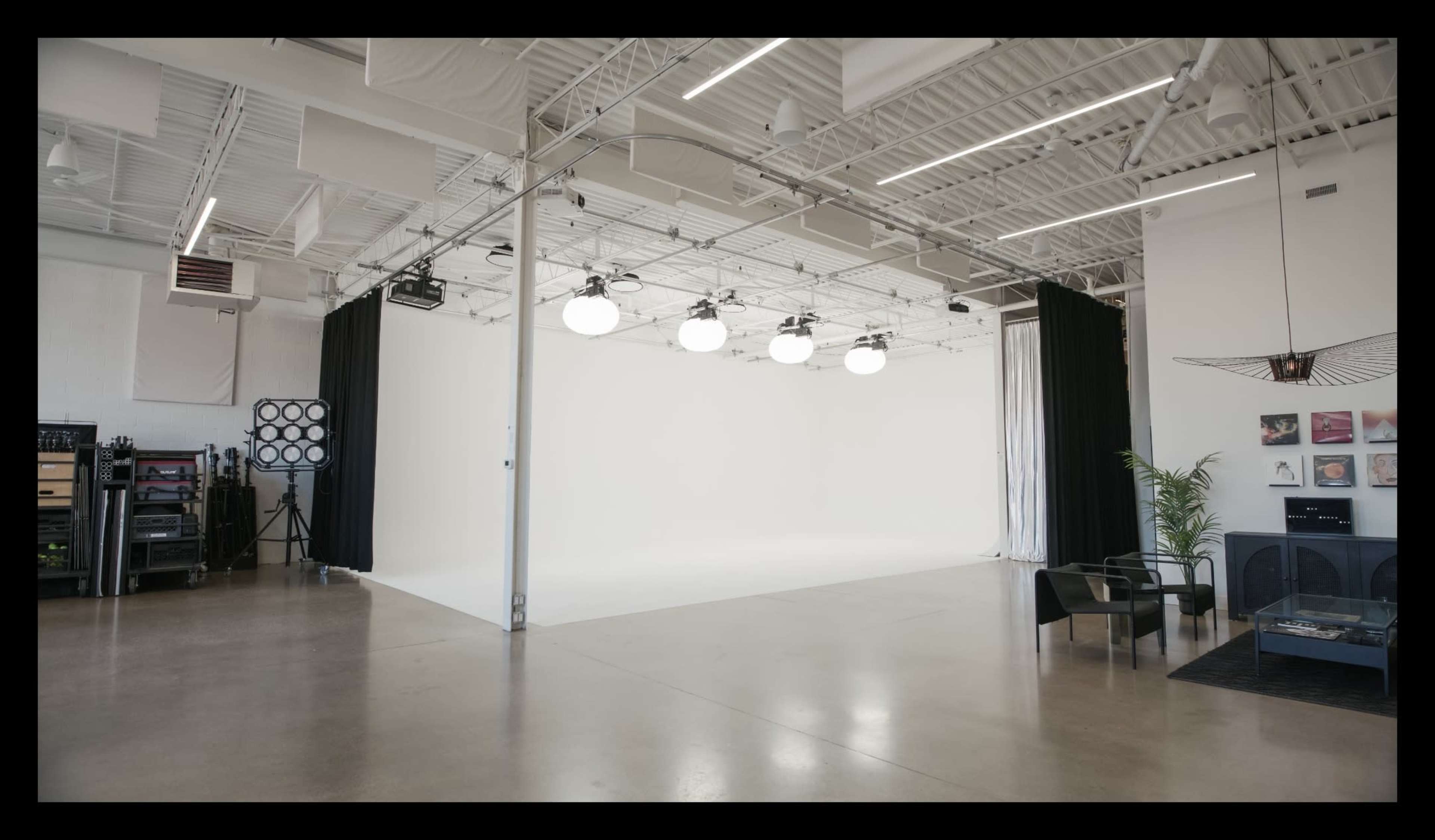 The image shows a spacious, well-lit studio with a white backdrop, overhead lighting, and minimalist furniture.