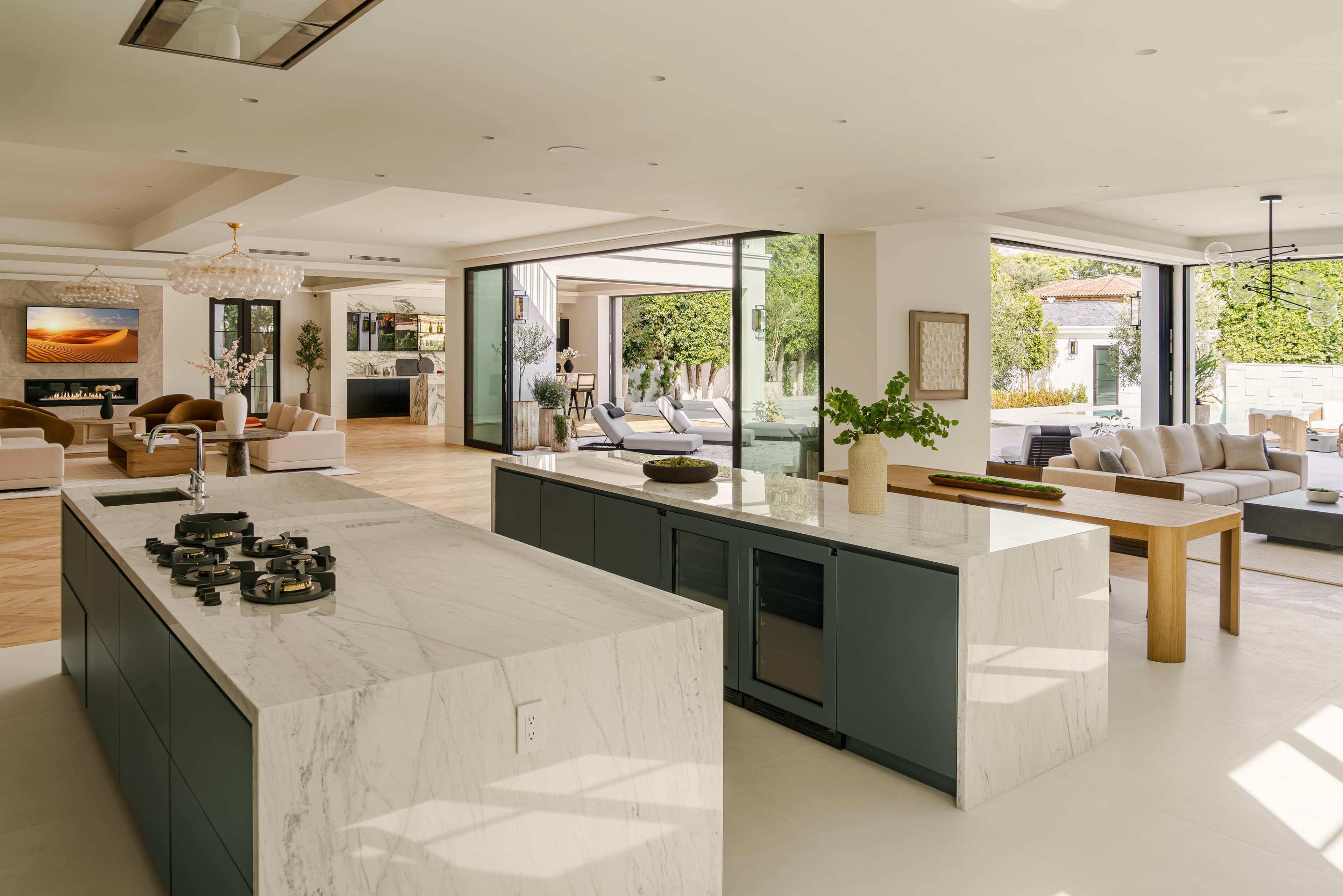 The image shows a modern open-concept kitchen with a marble island, integrated appliances, and large windows connecting to a living area and outdoor space.