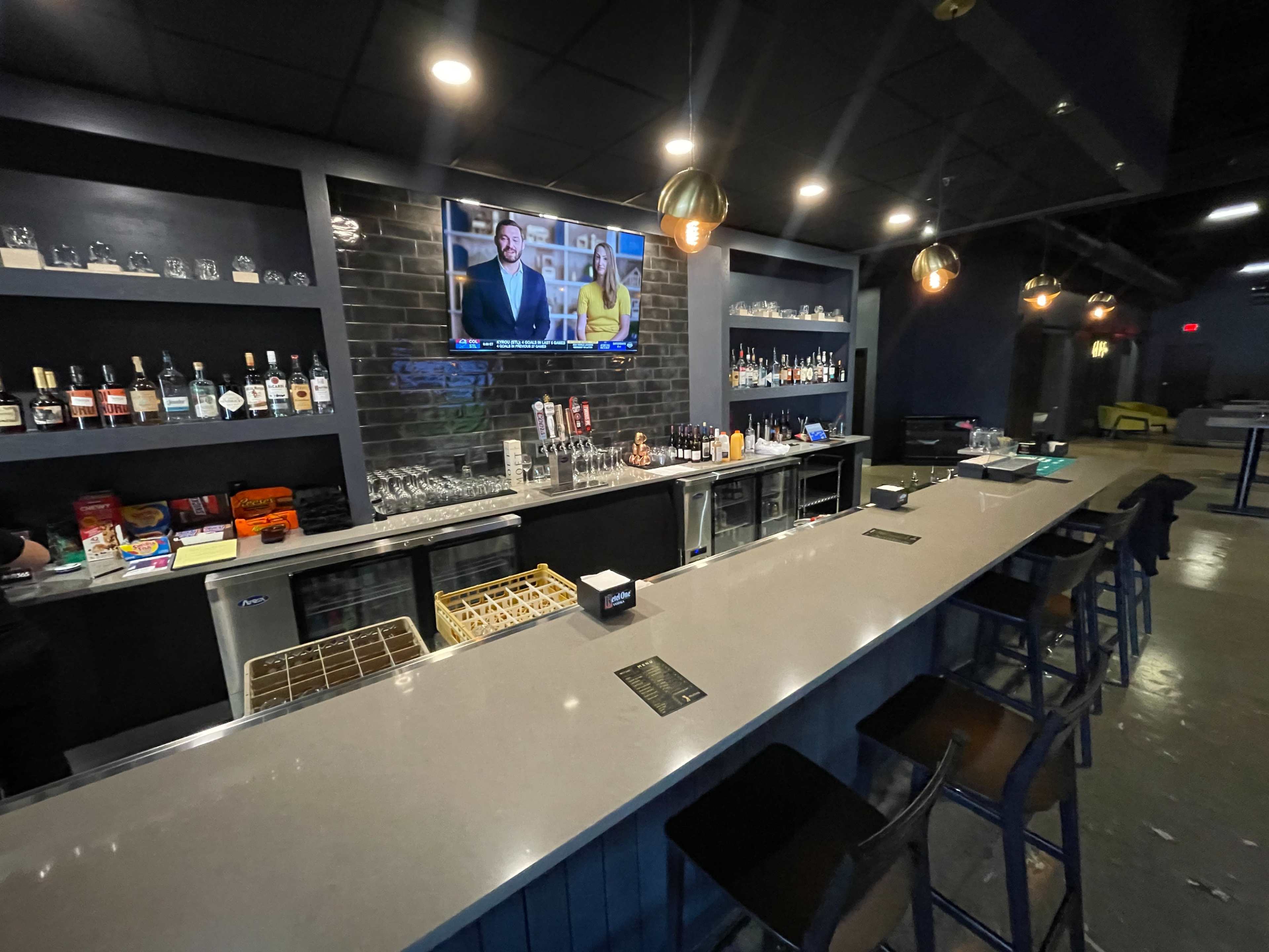 The image shows a modern bar with a long countertop, shelves lined with various bottles, and a television mounted on the wall displaying content.