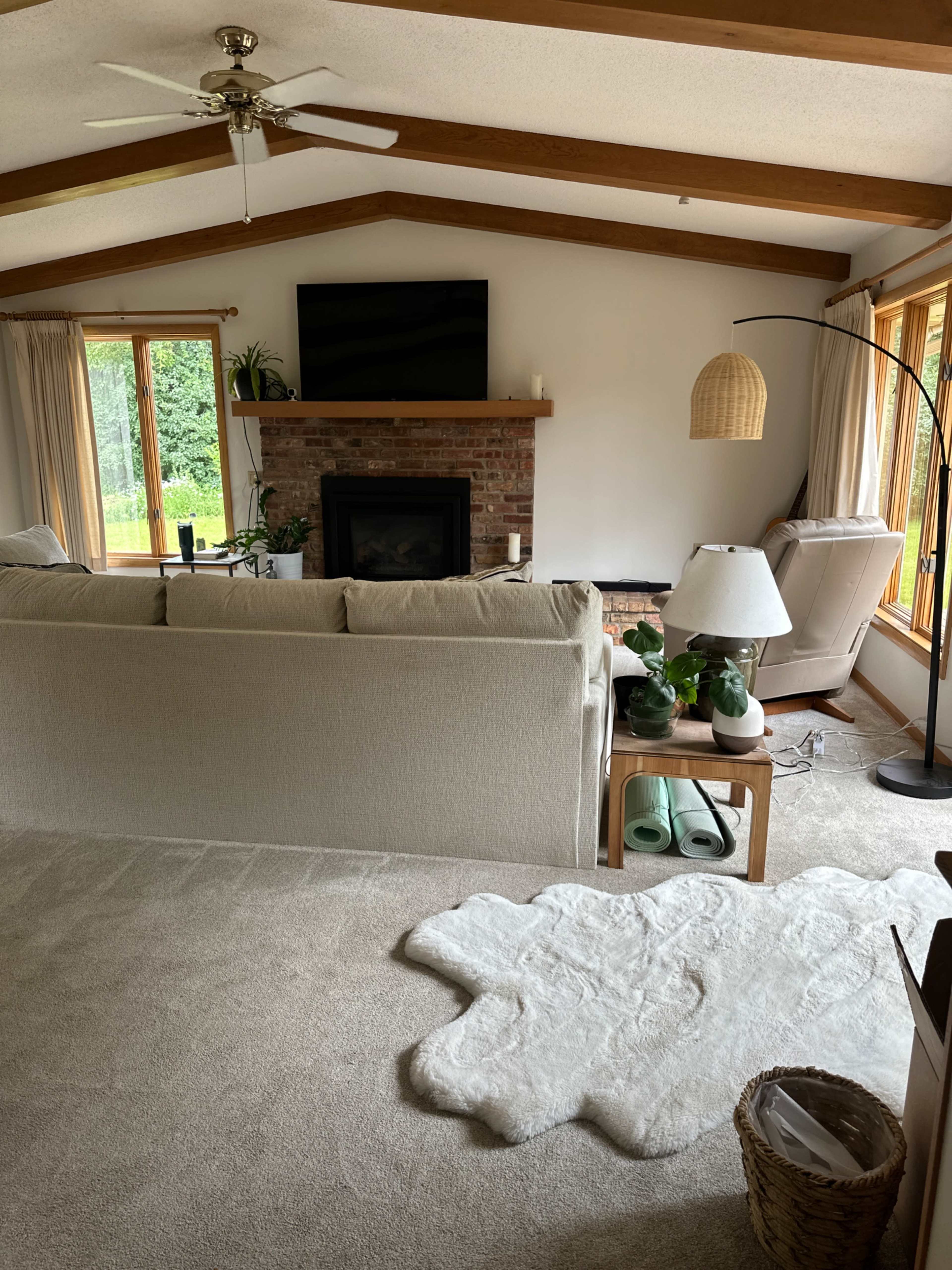 The image shows a cozy living room featuring a beige couch, a brick fireplace with a television above it, and large windows allowing natural light to enter.