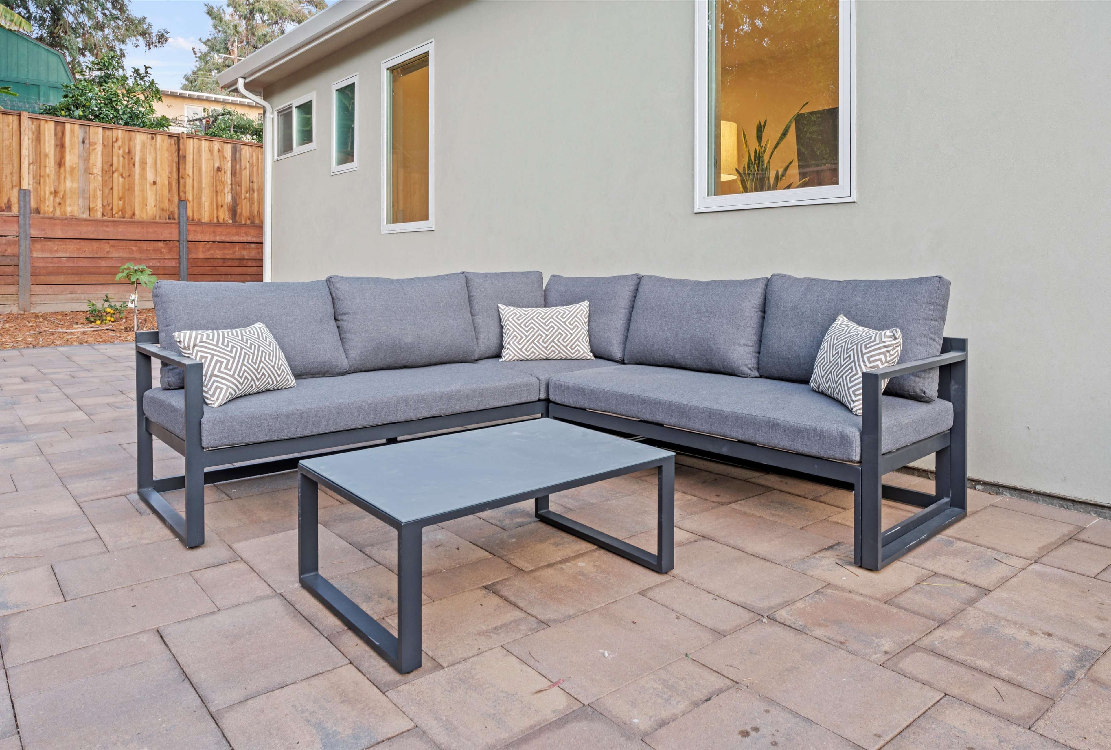 A gray sectional outdoor couch with two decorative pillows and a matching coffee table sits on a paved patio beside a wooden fence and green shrubs.