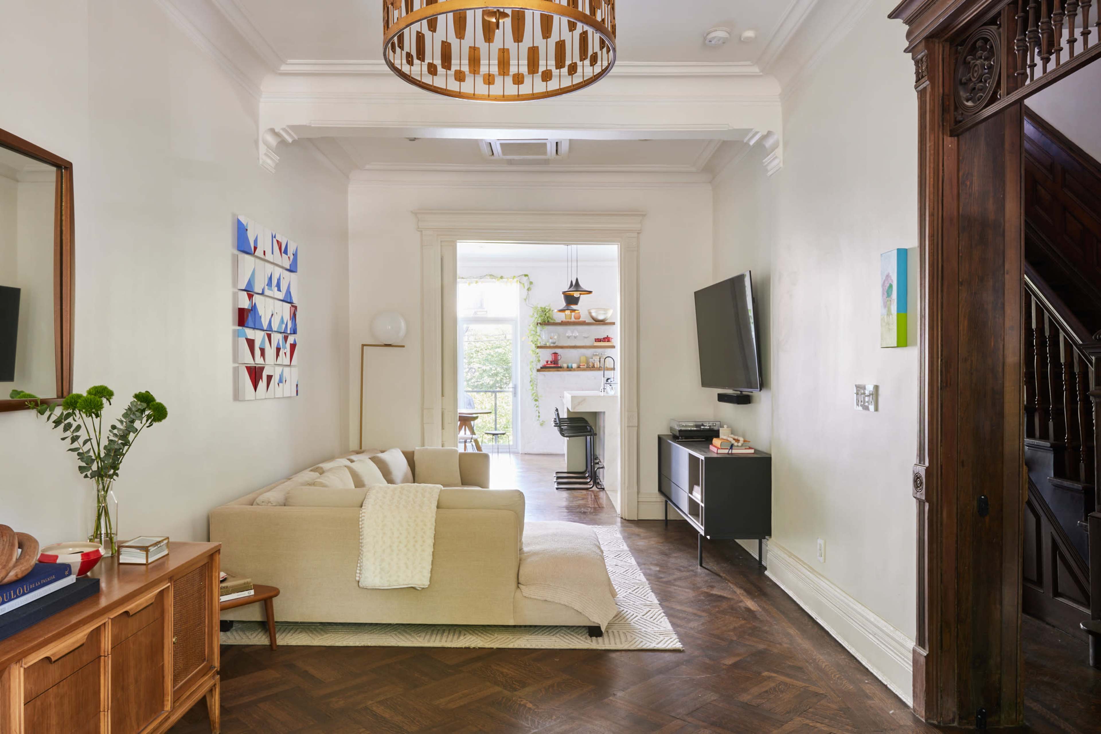 A well-lit living room featuring a beige sofa, a wooden cabinet, and a flat-screen TV, with an adjoining view of a kitchen area and large windows.