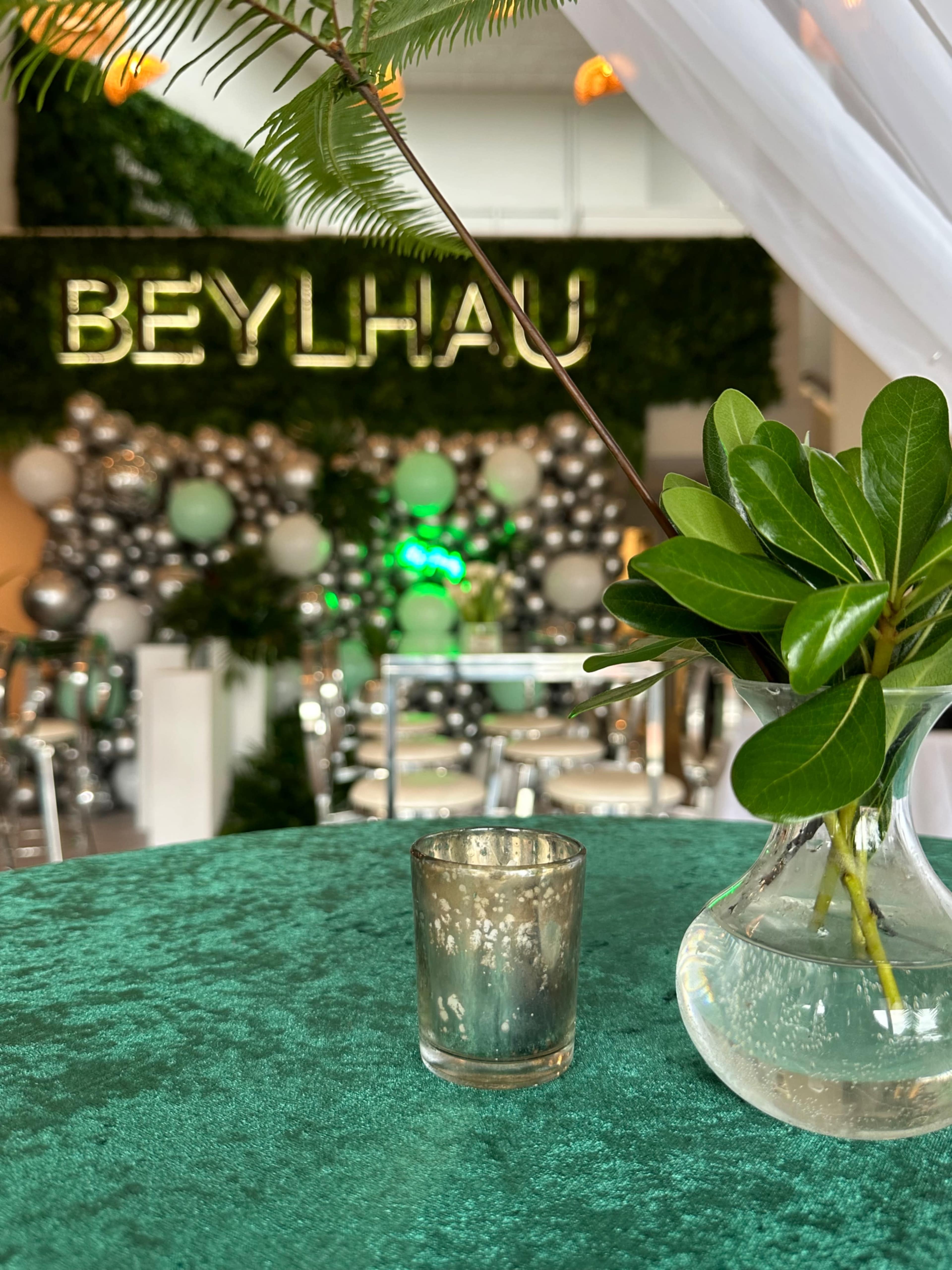 A table with a green tablecloth features a small vase of leaves and a decorative candle holder in front of a backdrop with the name "BEYLHAU" and a balloon arrangement.