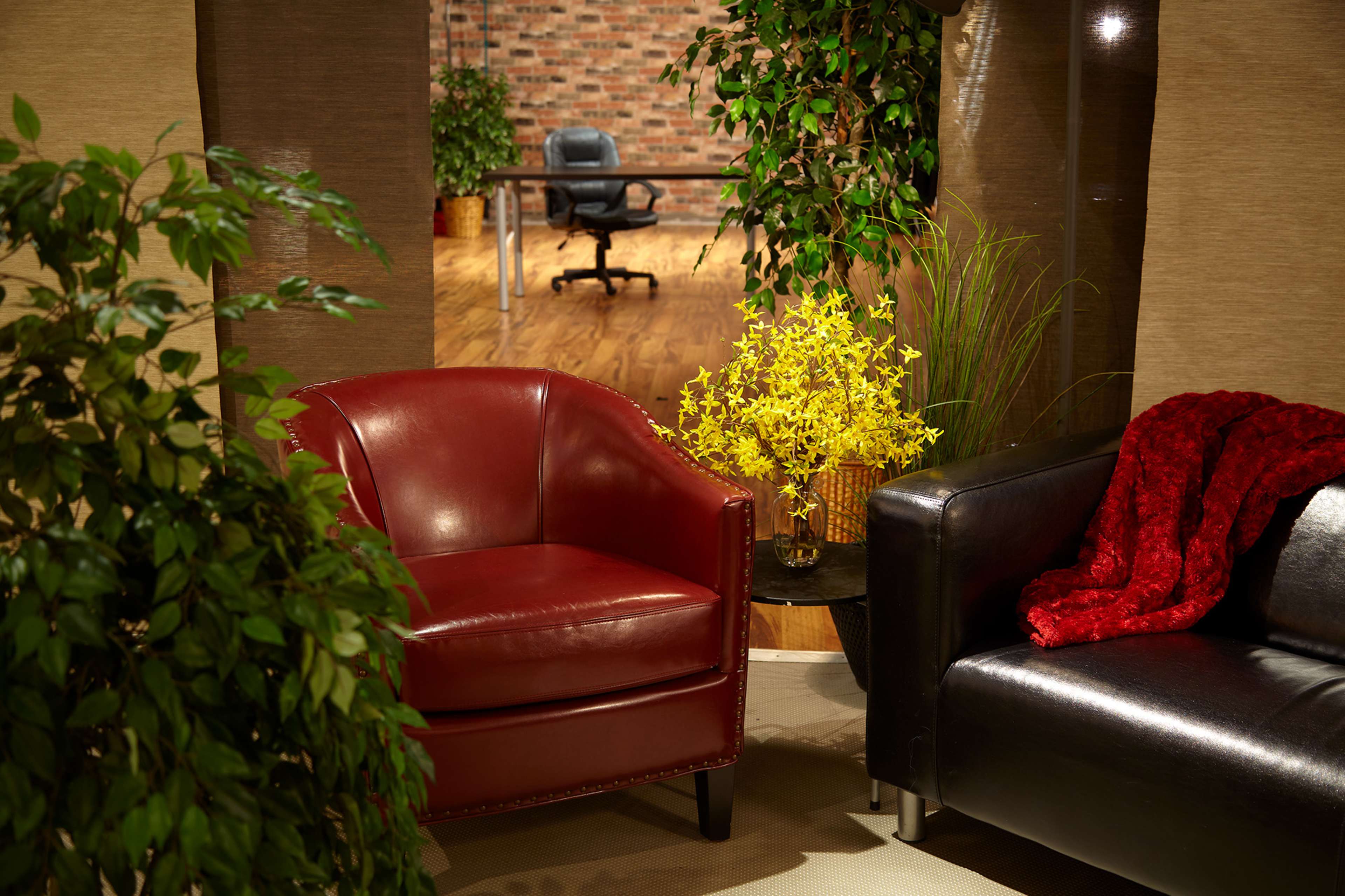 A cozy indoor space featuring a red armchair, a black sofa, a small table with a yellow flower arrangement, and a desk visible in the background.