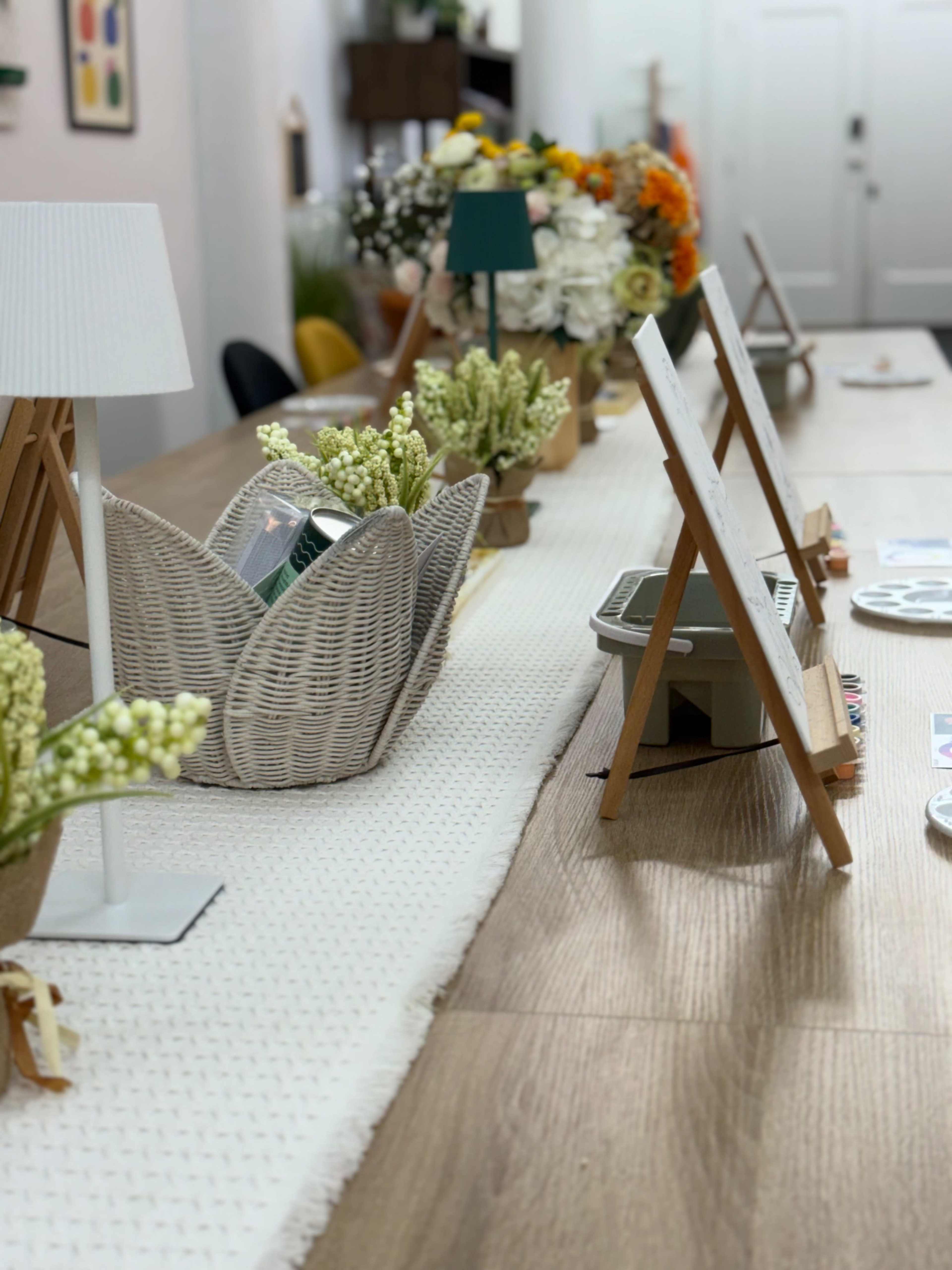 The image shows a long wooden table set with decorative elements, including flower arrangements, baskets, and small easels displaying items.