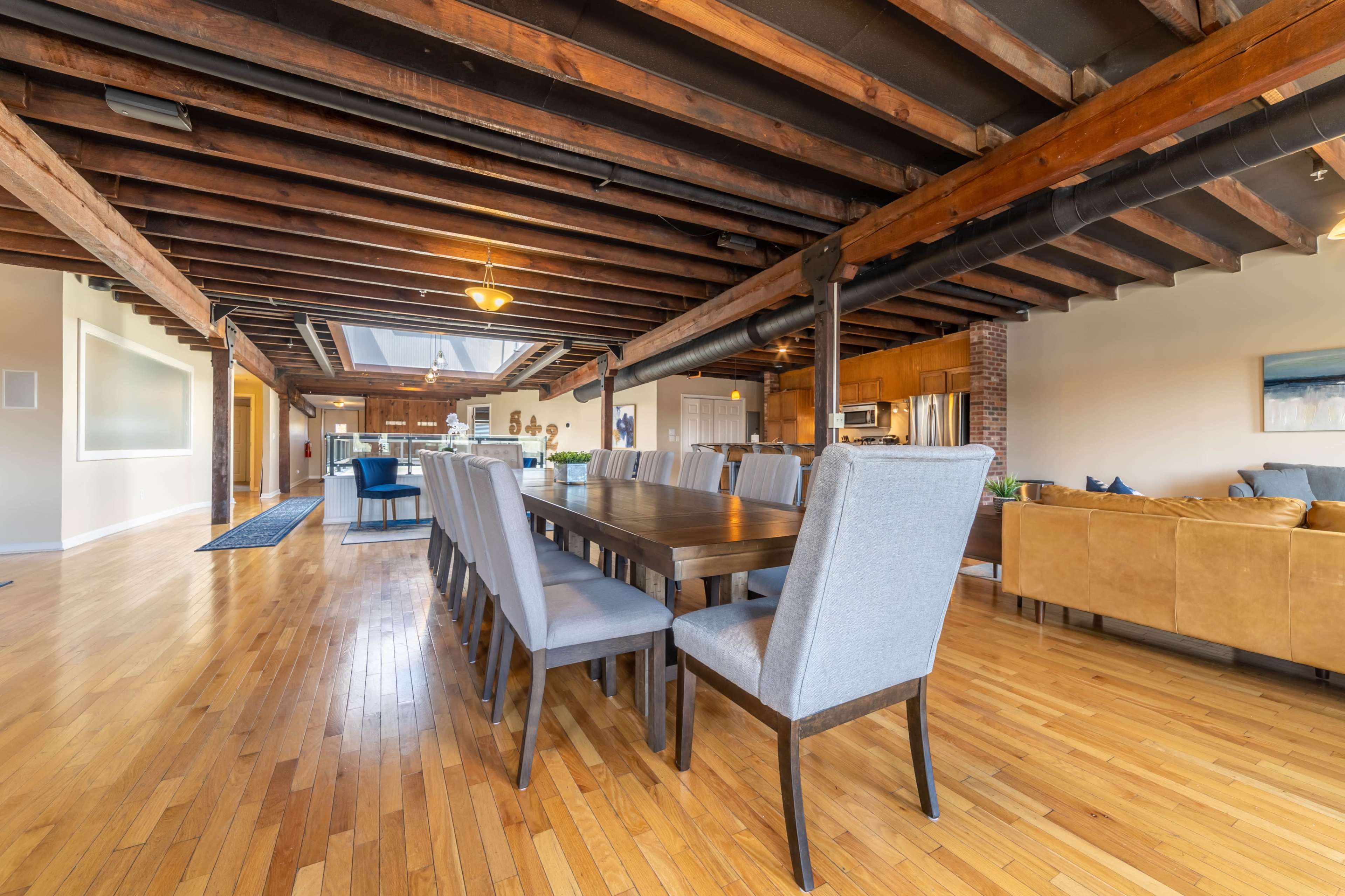 A spacious dining area featuring a long wooden table surrounded by gray upholstered chairs, with exposed wooden beams overhead.