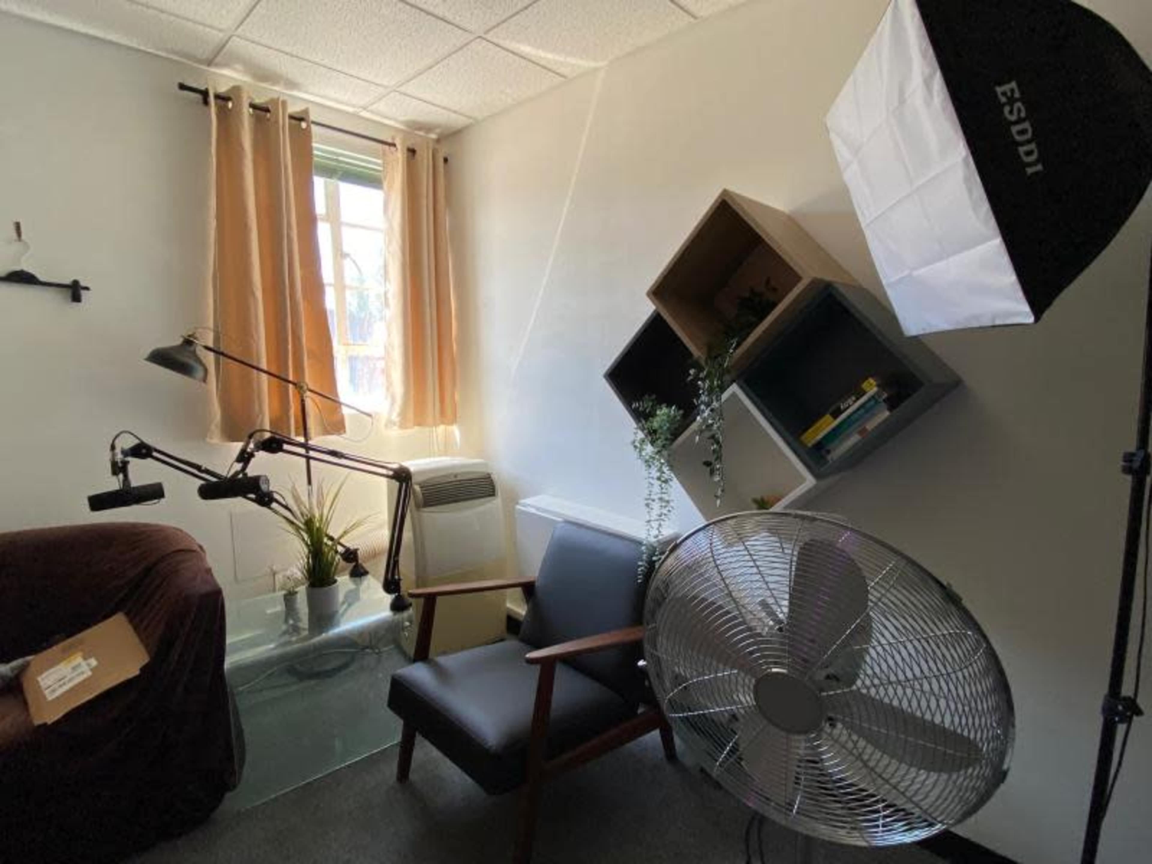 A small room with a fan, a chair, a glass table, a window with blinds, and shelves mounted on the wall.