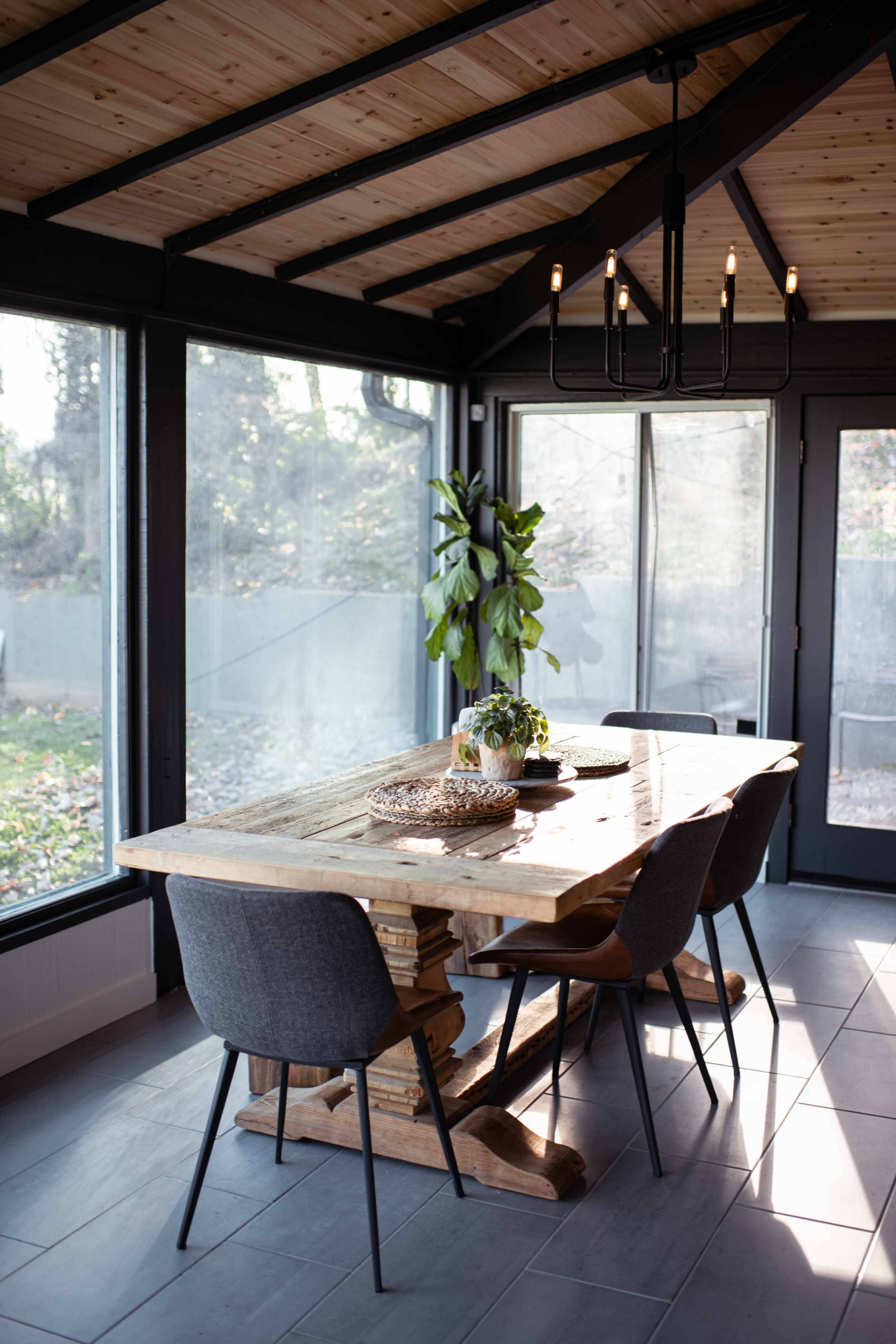 A wooden dining table with black chairs is set in a sunlit room featuring large windows and a wooden ceiling.