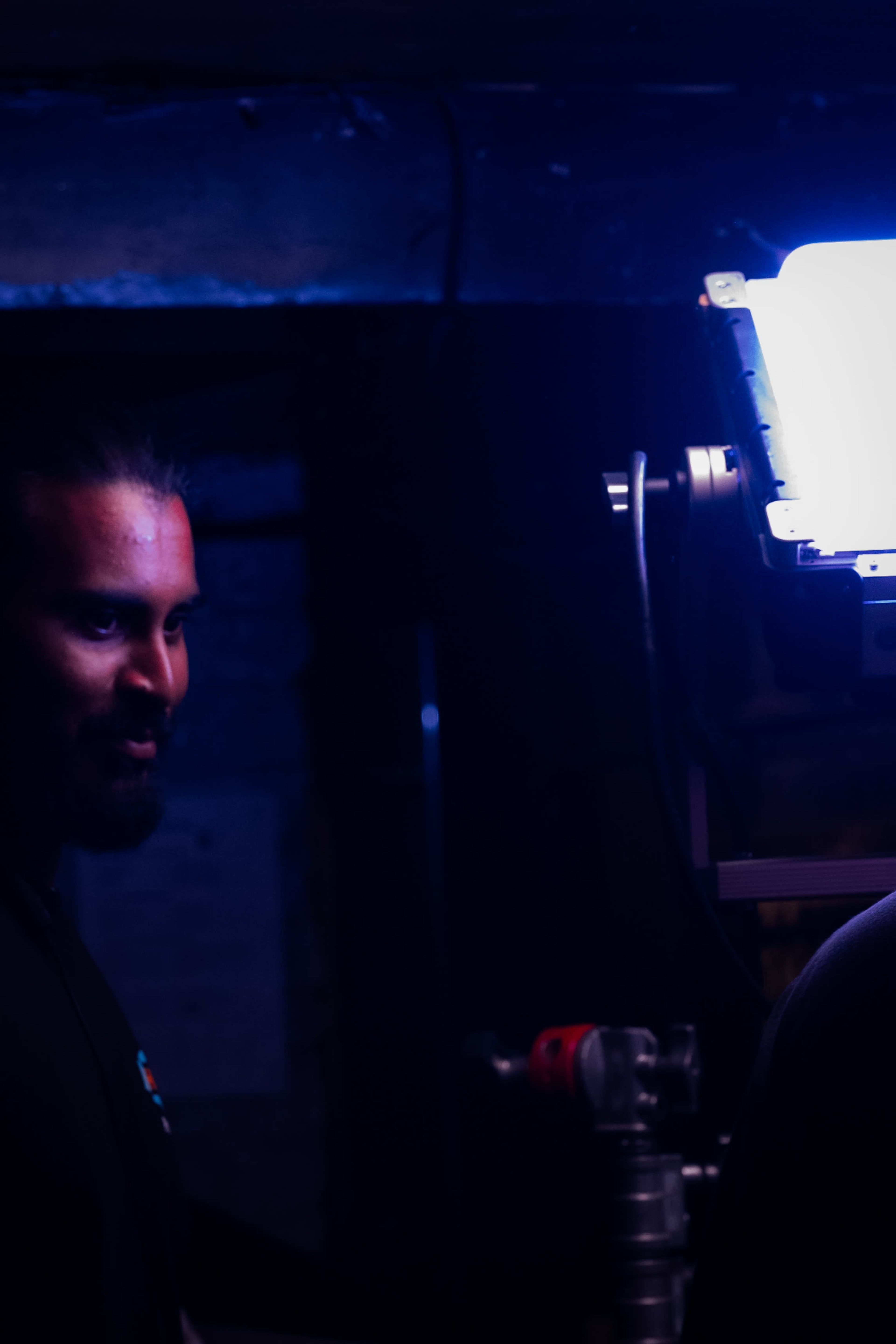 Two individuals are standing in a dimly lit space, with one person illuminated by a bright light source.