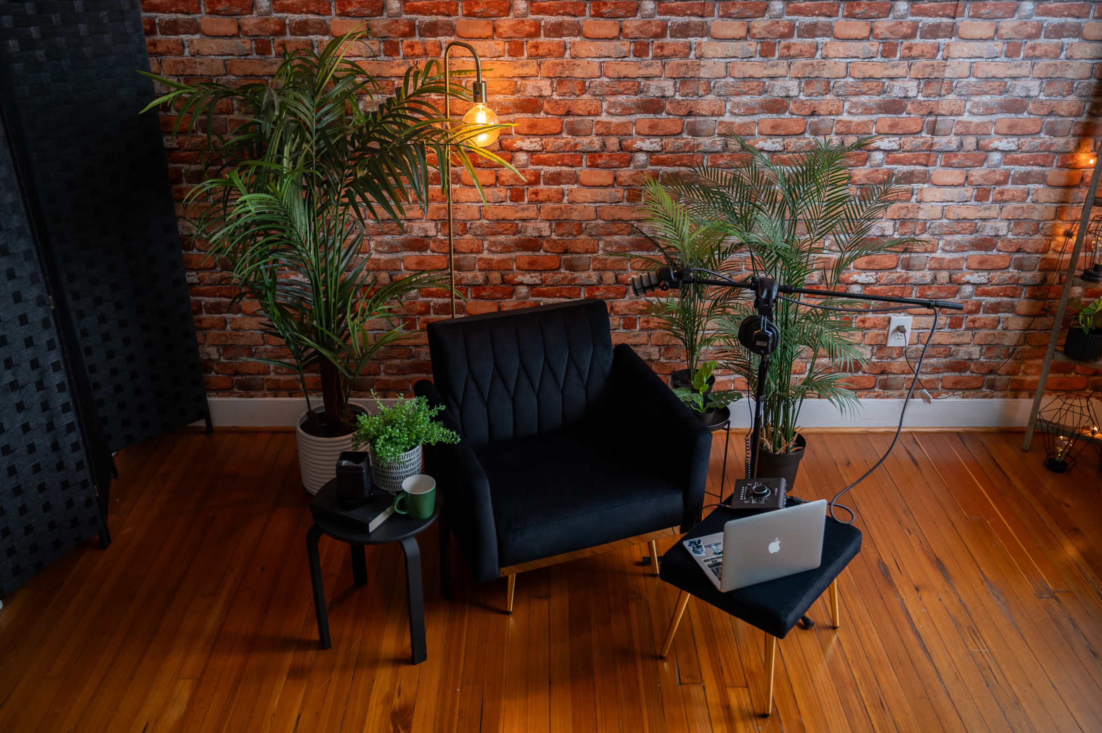A cozy seating area features a black armchair beside a small table with a laptop and a microphone, set against a brick wall and surrounded by potted plants.