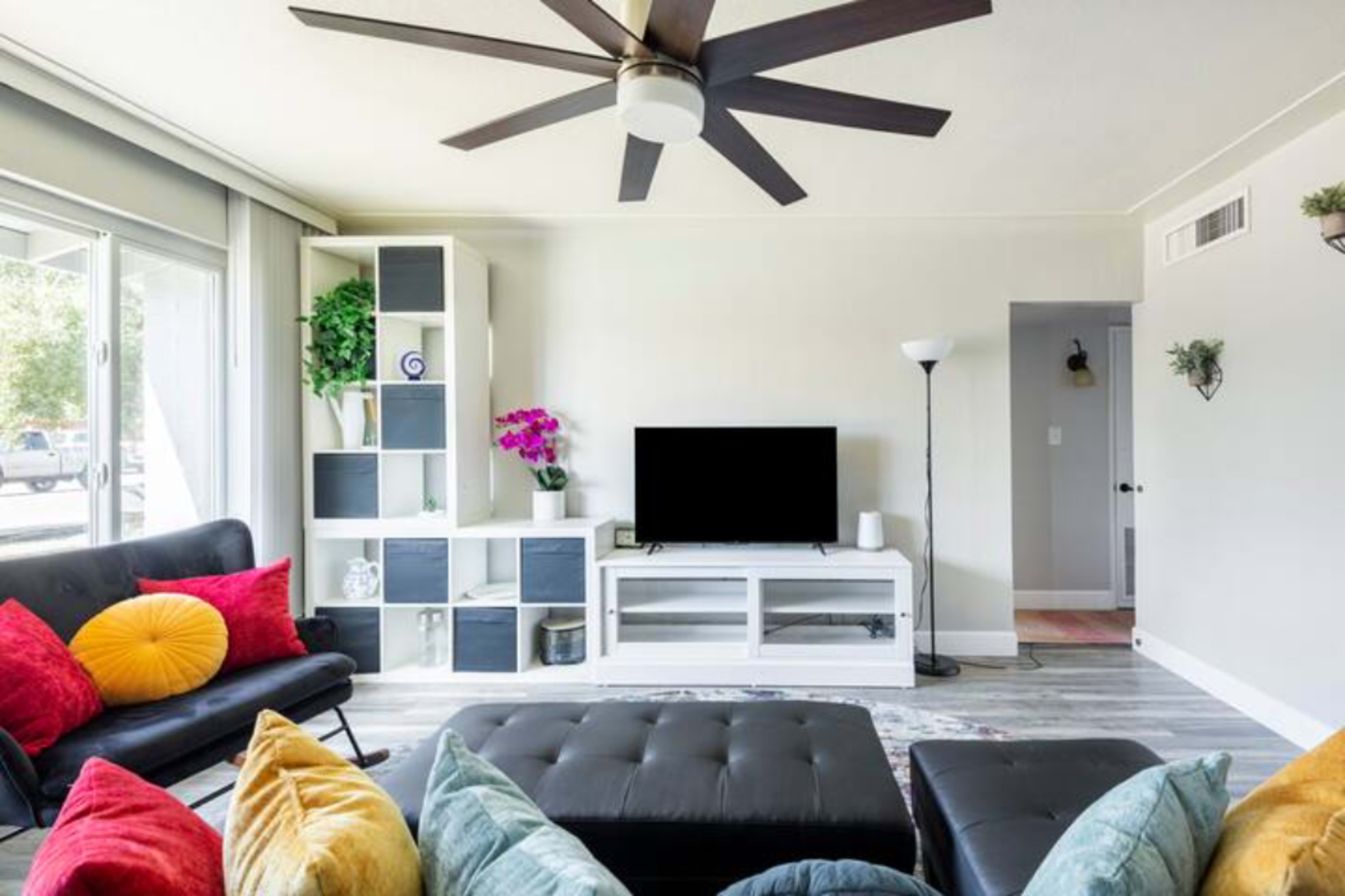 The living room features a modern design with a black ceiling fan, a dark sofa, colorful cushions, a sleek entertainment unit, and large windows allowing natural light.