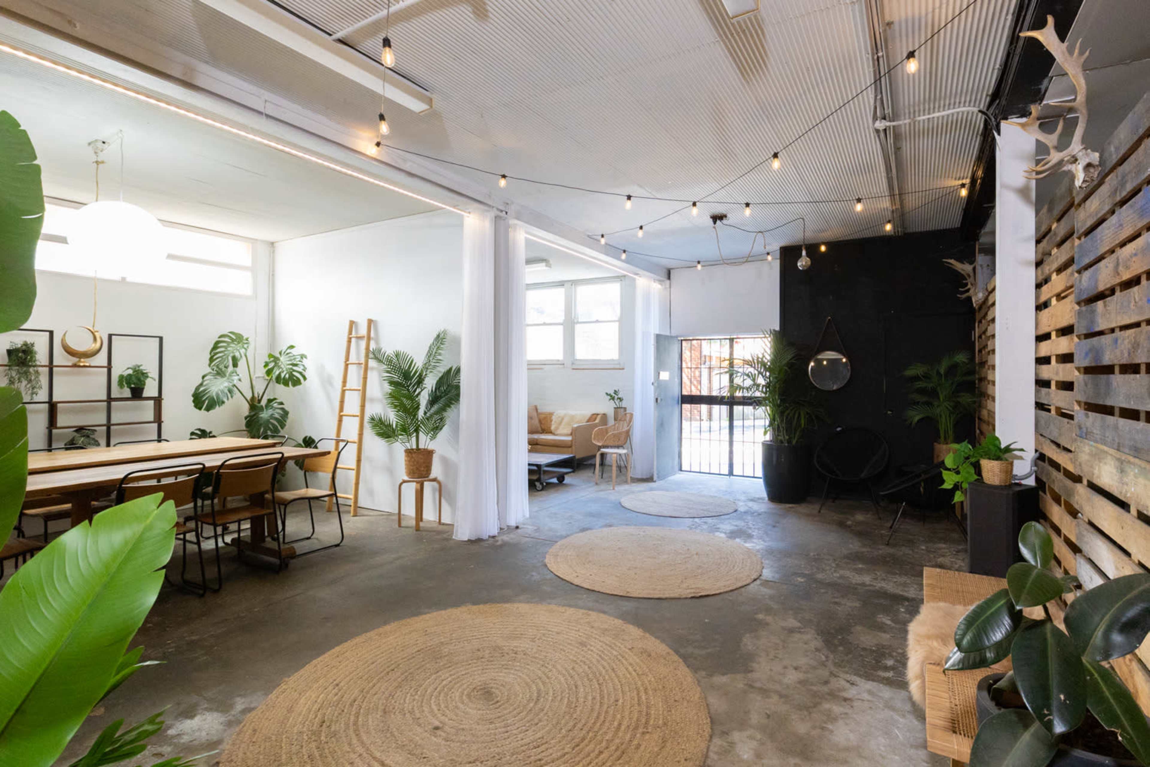 The image shows a modern, open space featuring a dining area with a long wooden table, a cozy sitting area, and various plants, illuminated by string lights and natural light from windows.