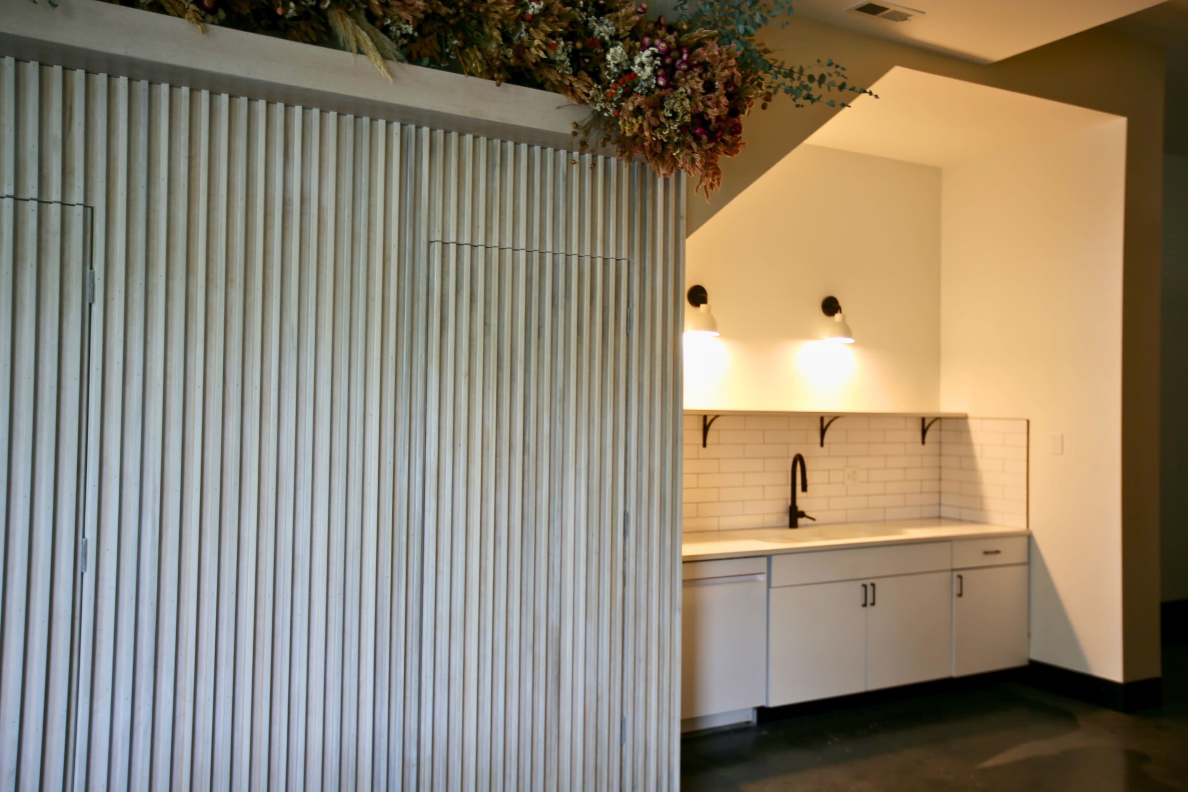 A modern kitchen area featuring a wooden paneled wall with a minimalist sink and countertop under wall-mounted lights.