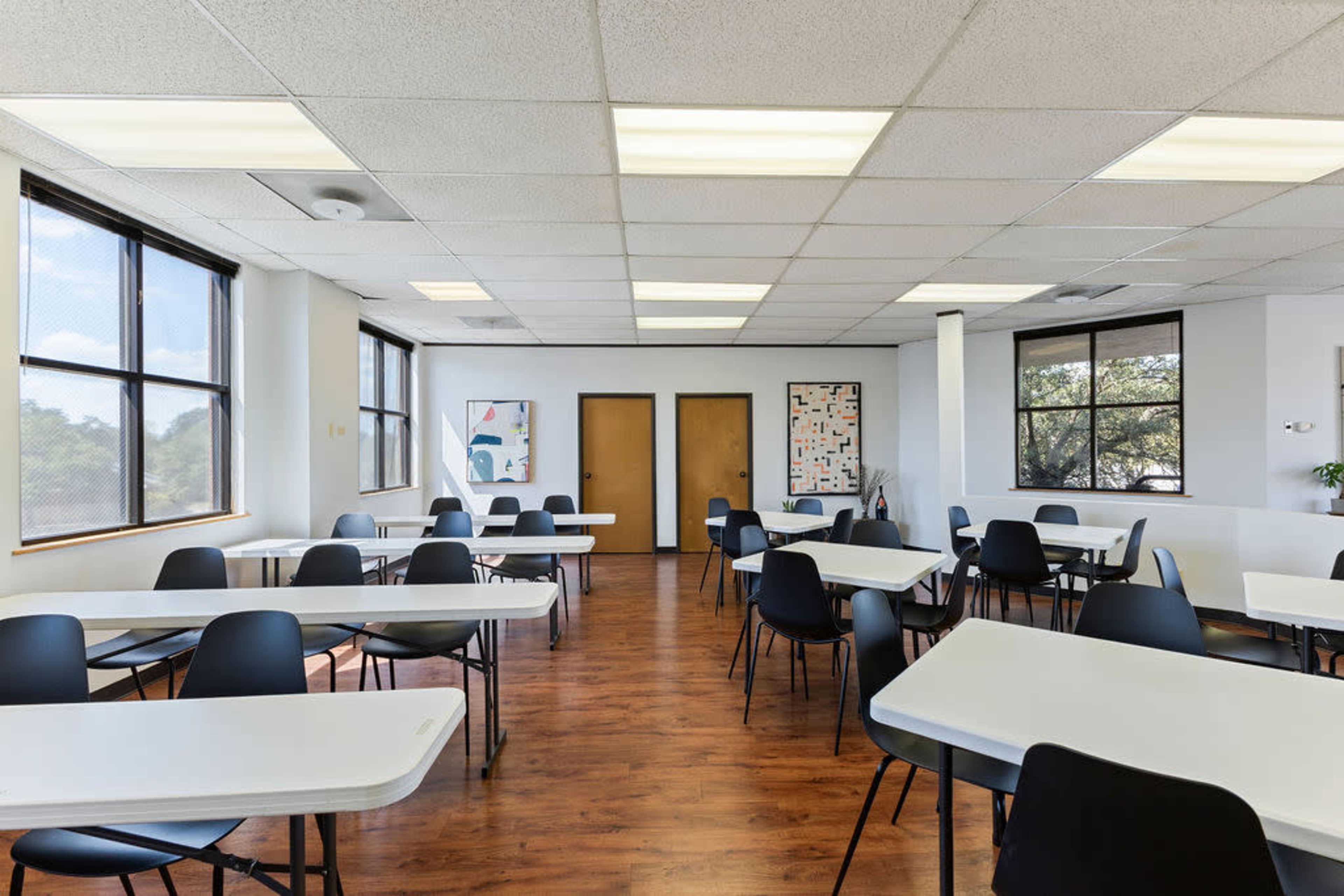 The image shows a spacious, well-lit classroom or meeting room with several tables and chairs arranged throughout the area.