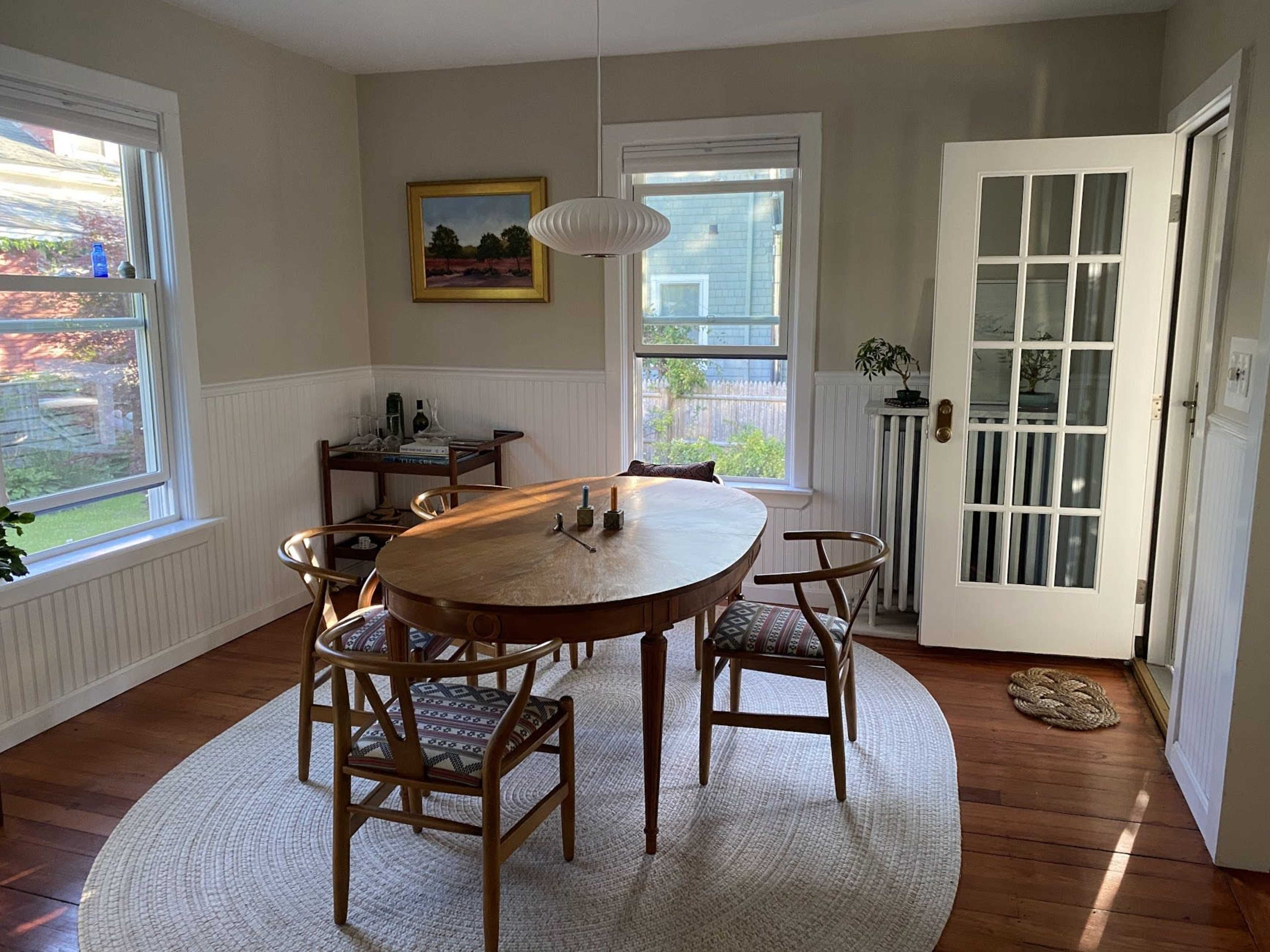 A round wooden dining table surrounded by four chairs is set in a light, airy dining room with large windows and a door leading outside.