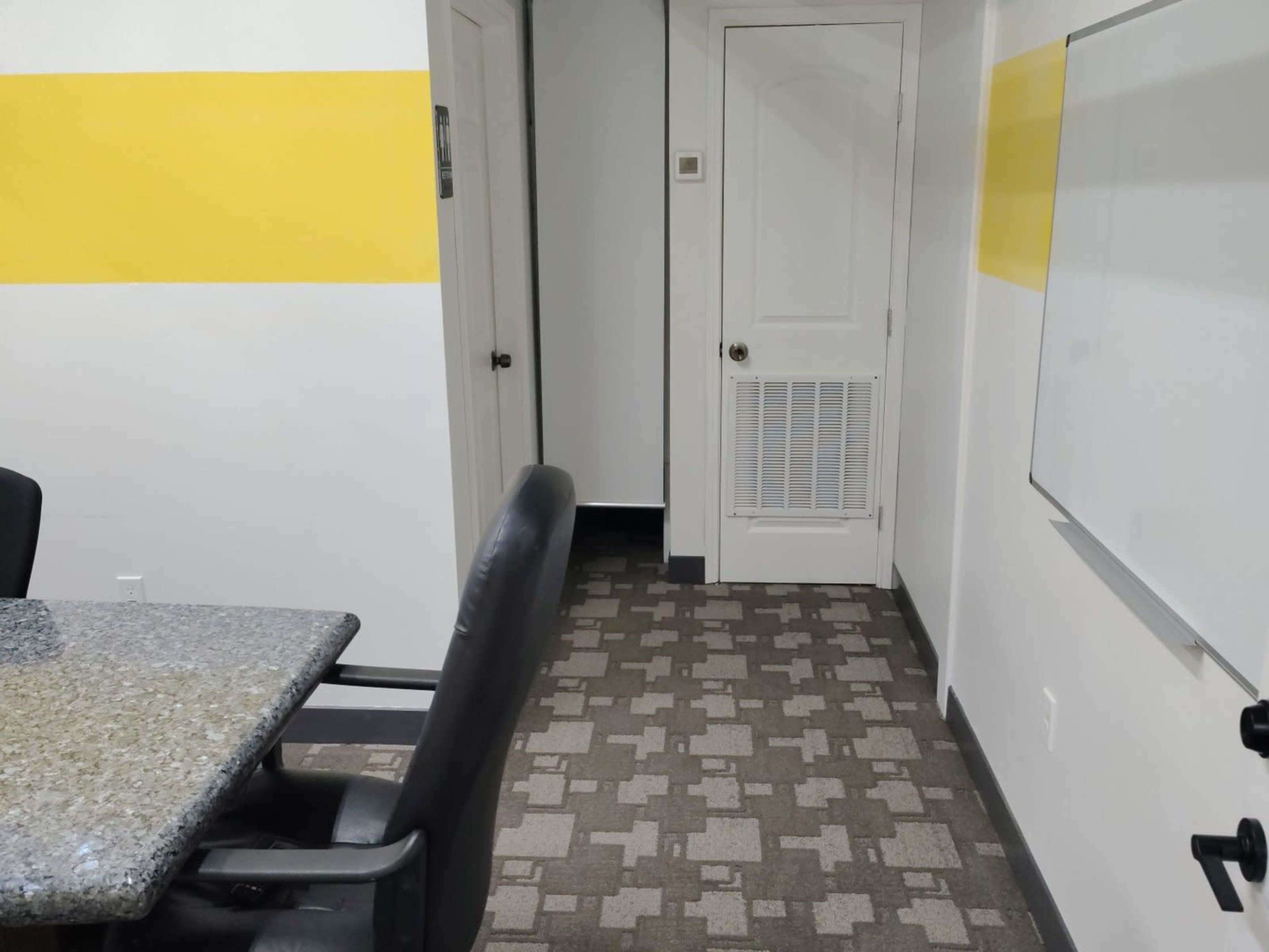 The image shows a meeting room with a granite table and black chairs, leading to a hallway with a door and a whiteboard on the wall.