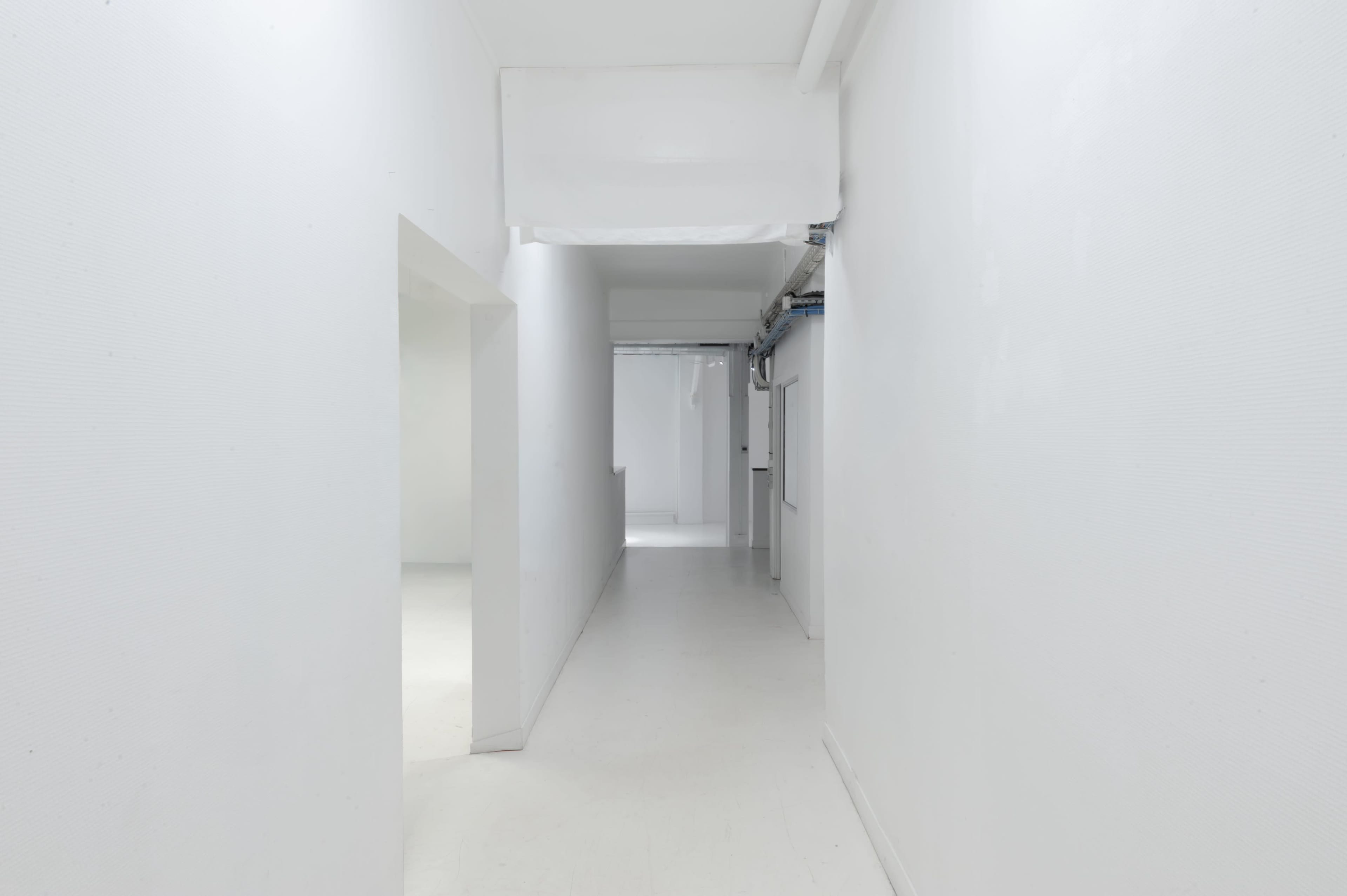The image shows a long, empty hallway with white walls and a white floor, leading to an open doorway at the end.