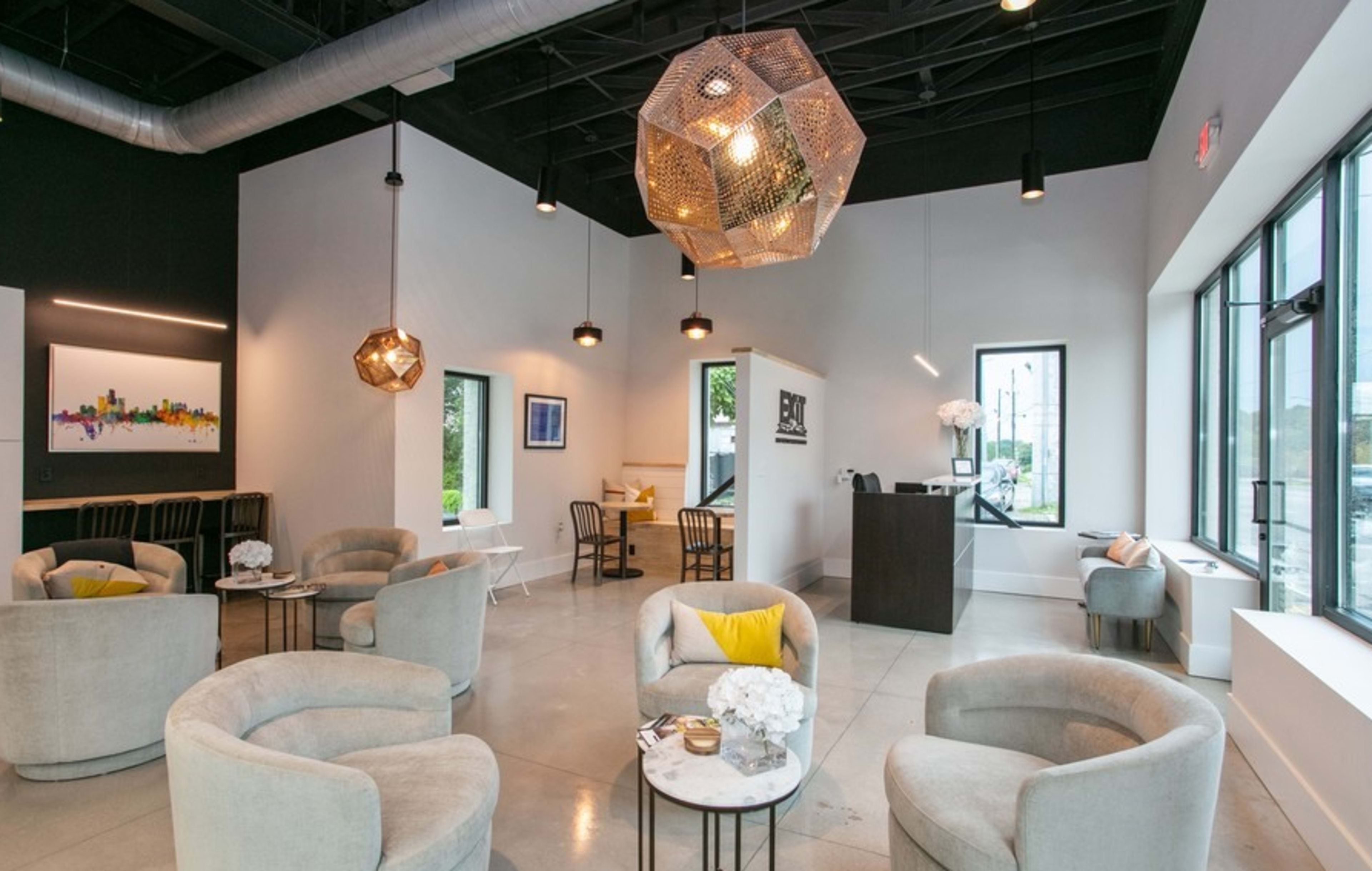A modern interior of a lounge area with gray upholstered chairs, geometric light fixtures, and large windows providing natural light.