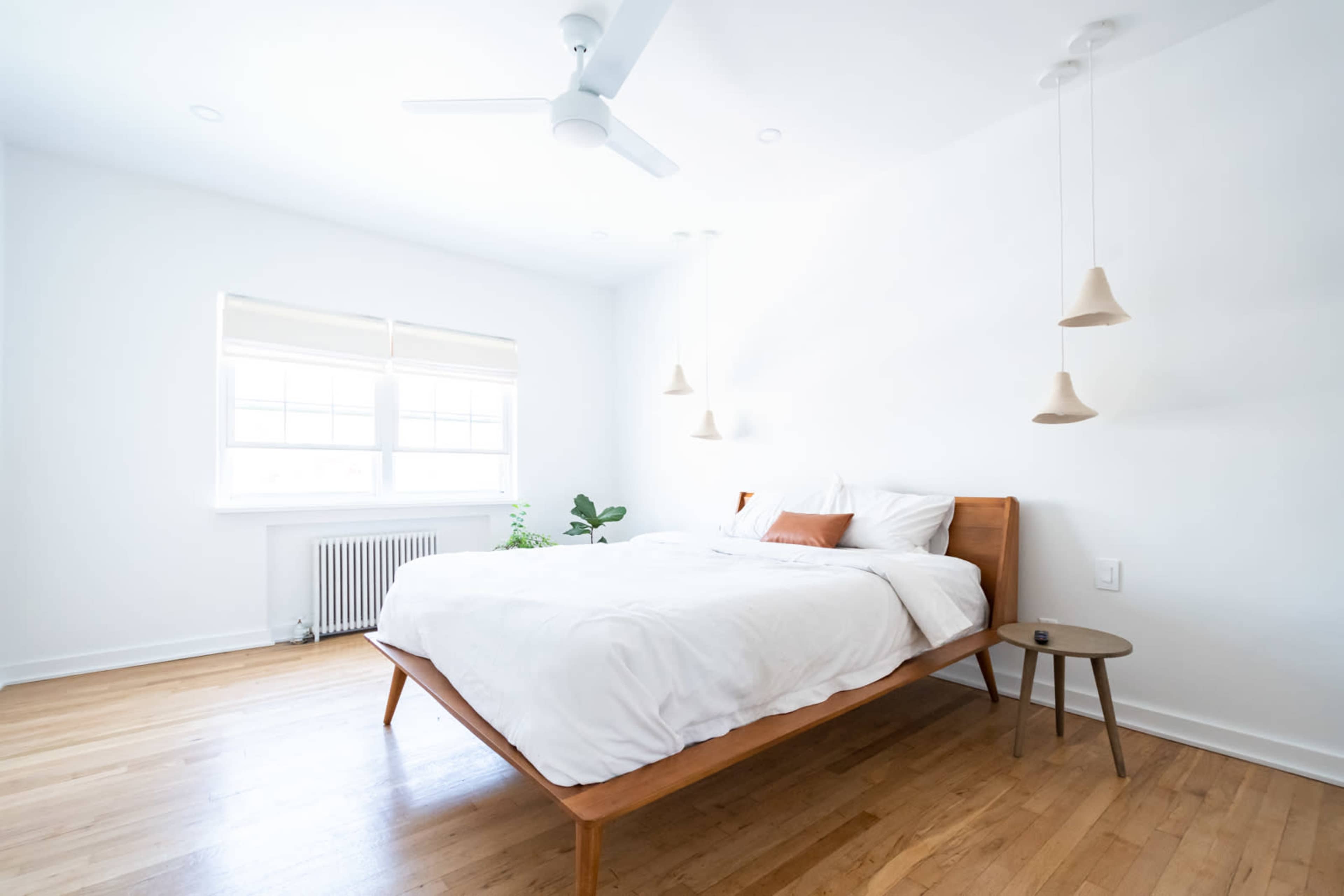A minimalist bedroom featuring a wooden bed with white bedding, a side table, and pendant lights, all set against white walls and hardwood flooring.