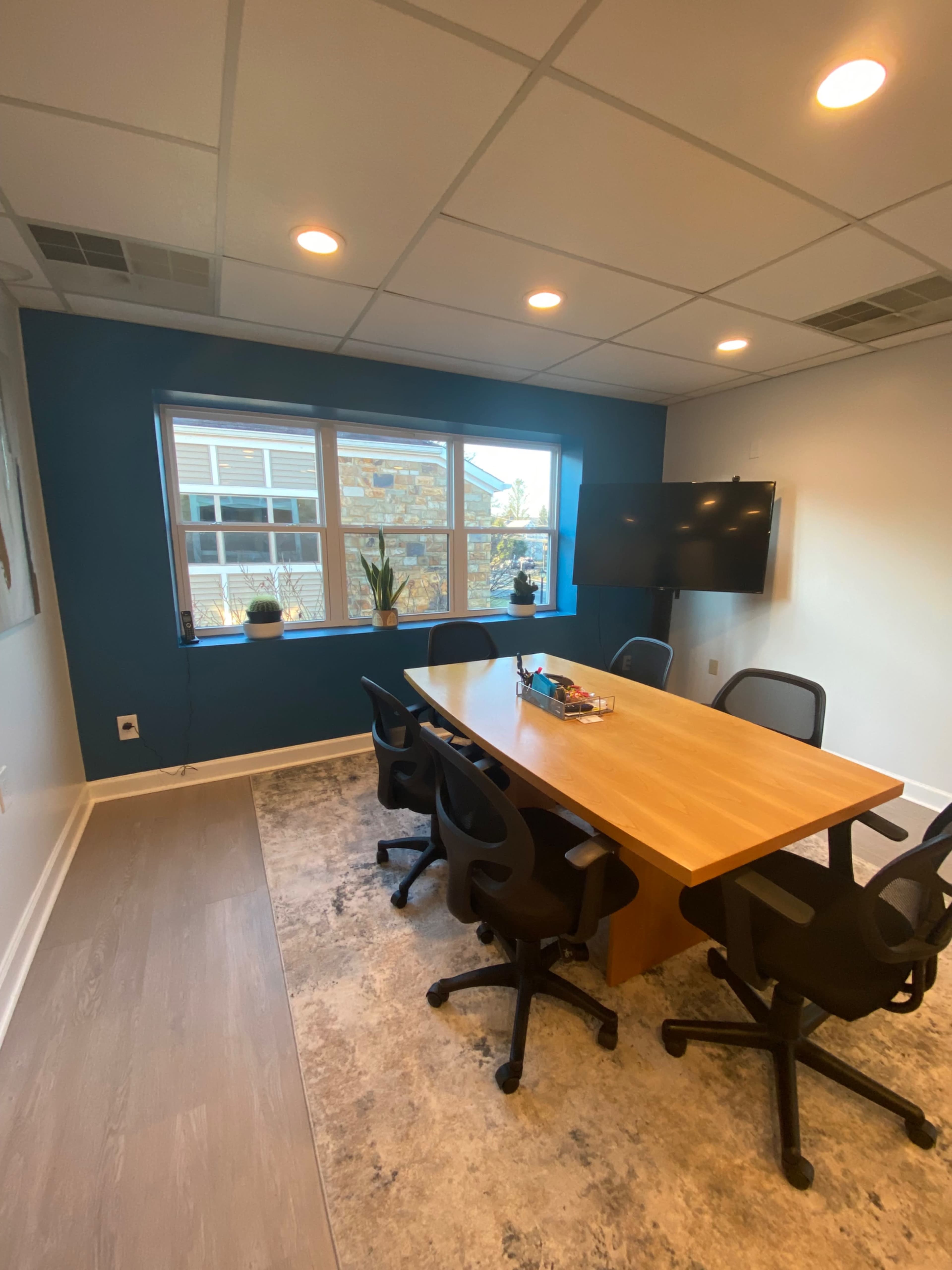 The image shows a conference room with a large wooden table surrounded by black chairs, a television mounted on the wall, and a window providing natural light.