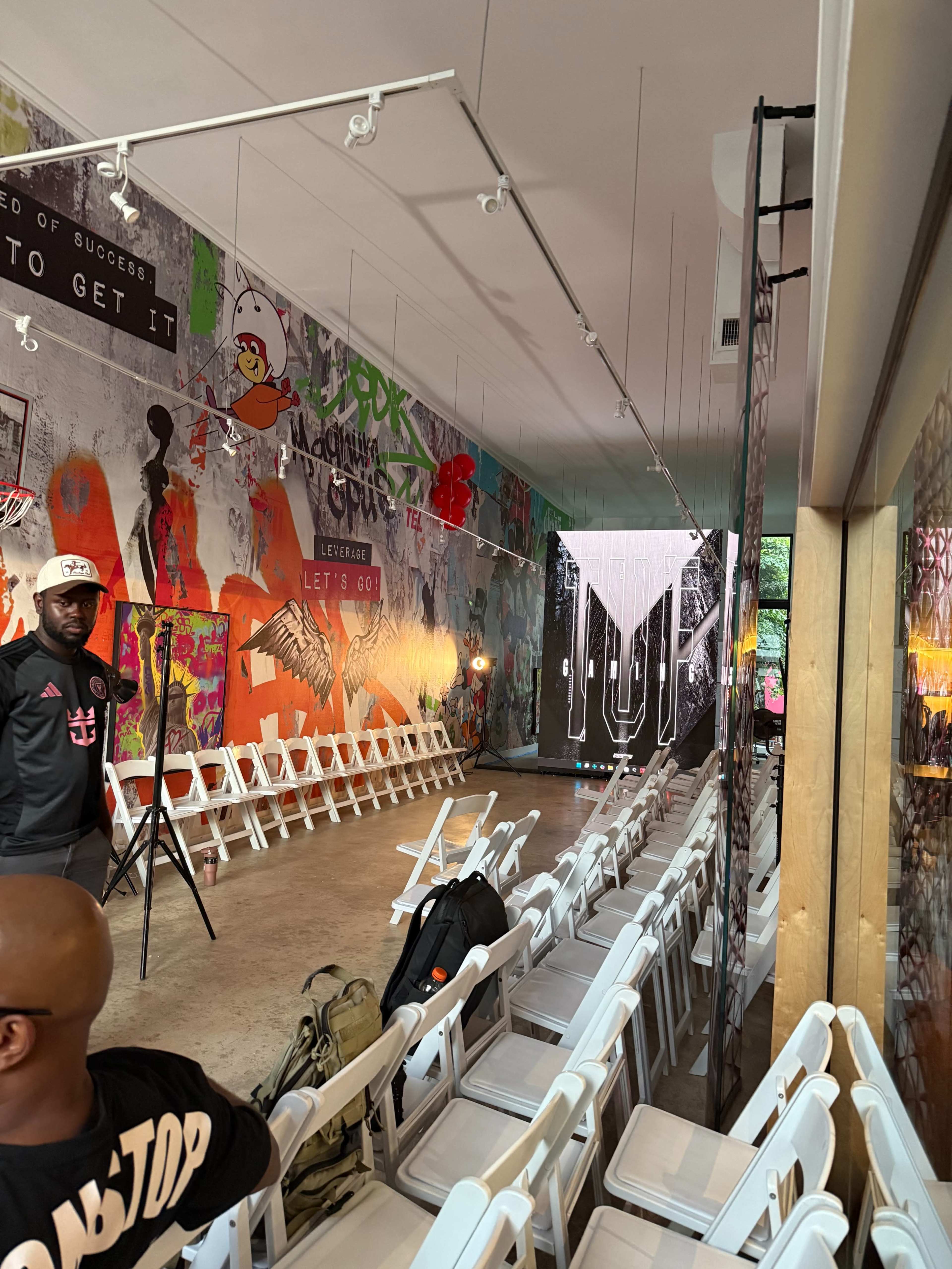 The image shows a colorful, graffiti-covered interior space set up for an event, with white chairs arranged in rows and a large decorative curtain at one end.